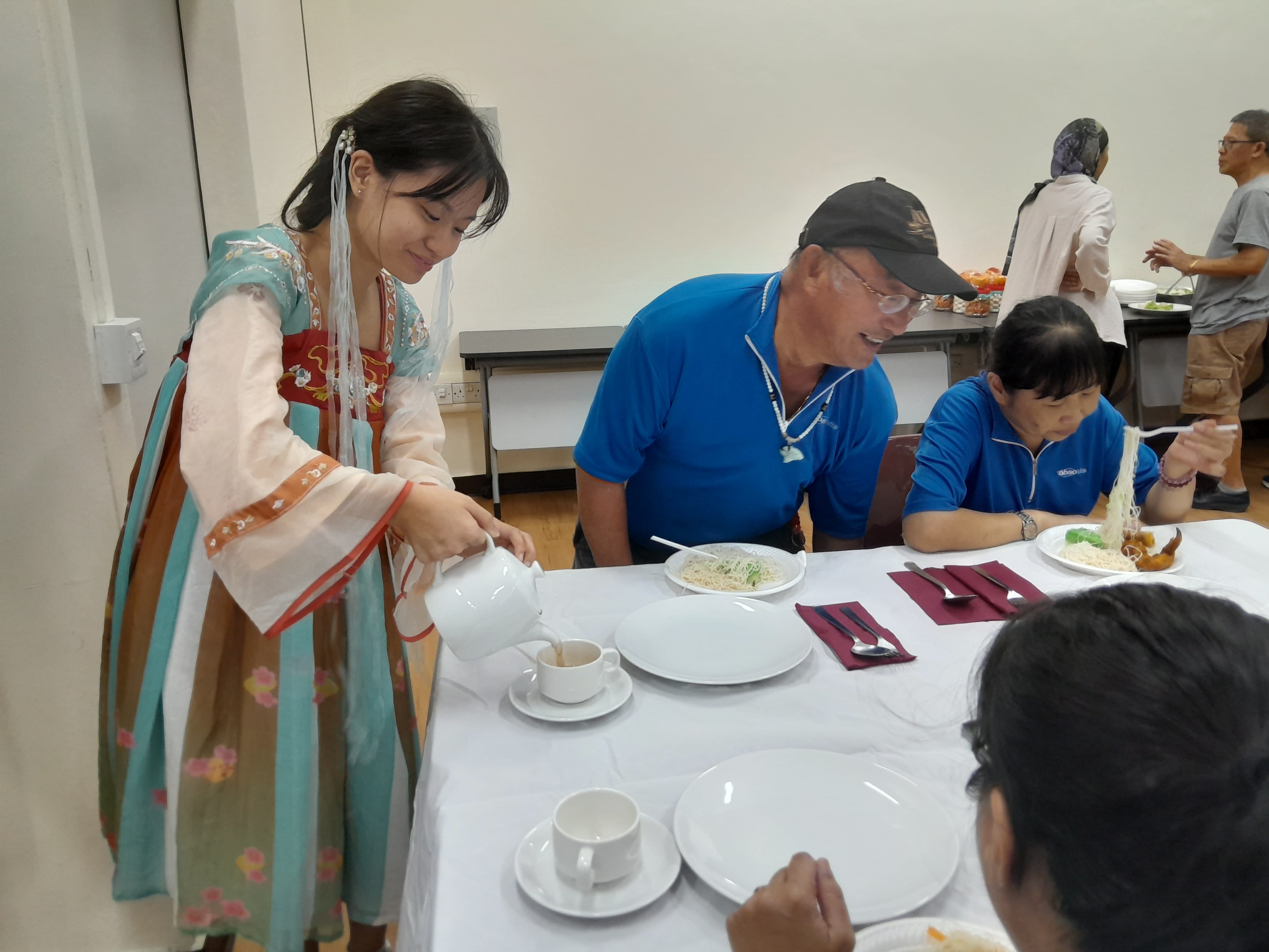 Woman in traditional Chinese dress pours tea for diners at a white-clothed table with noodle dishes.