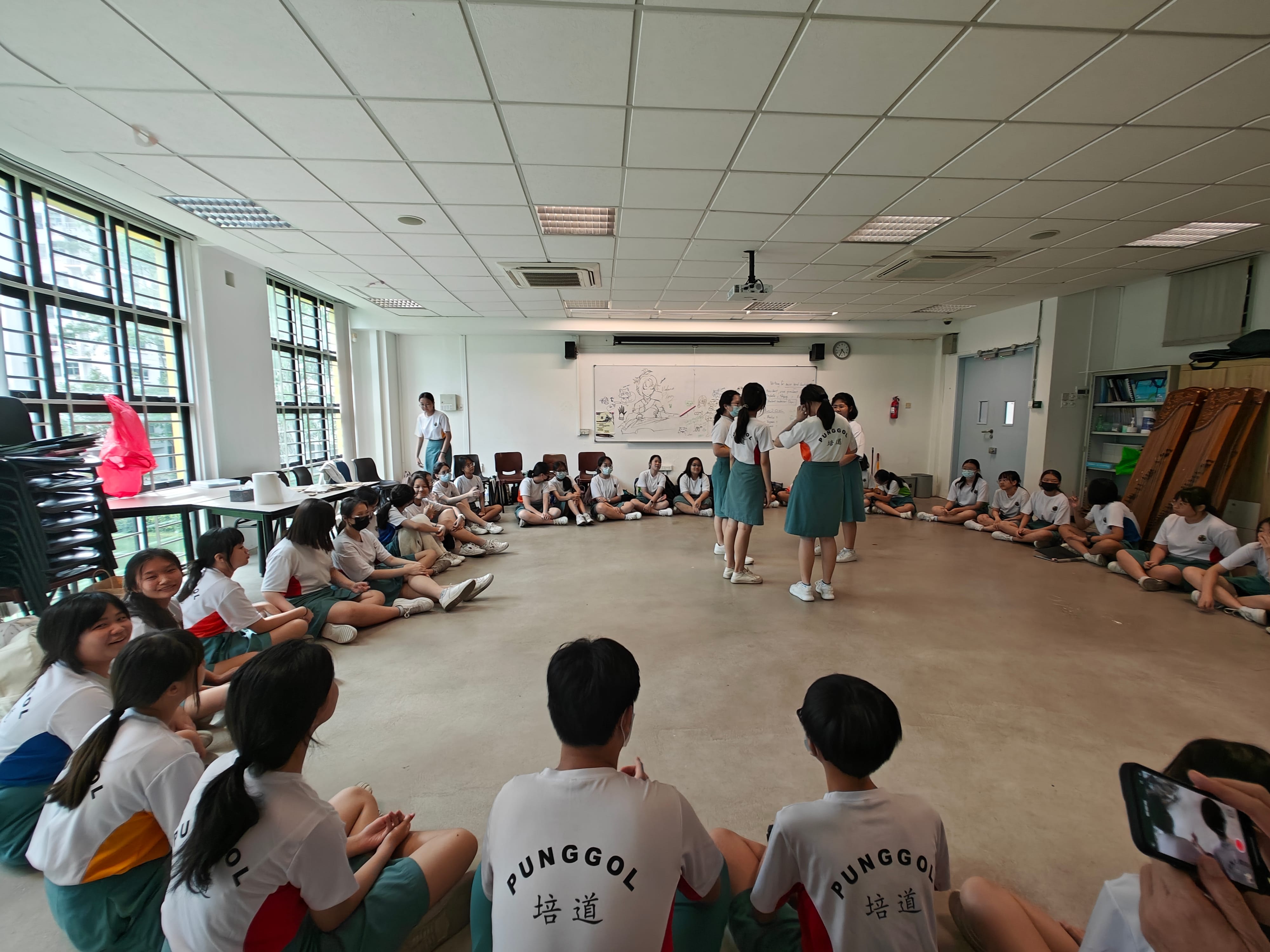 Students in Punggol school uniforms sit in a circle on the floor while two students stand in the center.