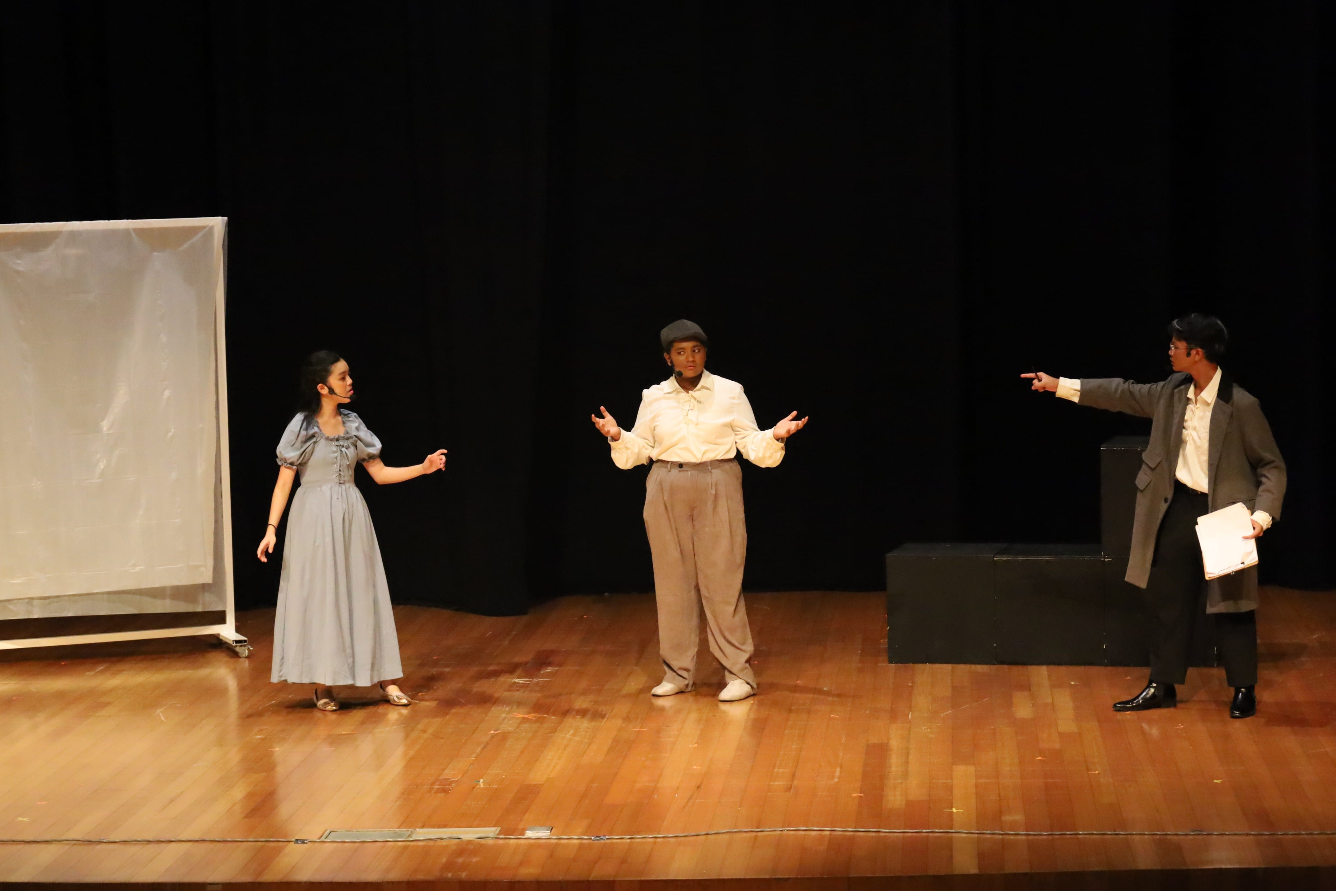 Three performers on a wooden stage, one in a blue dress, one in a white shirt gesturing, one in a gray suit pointing.