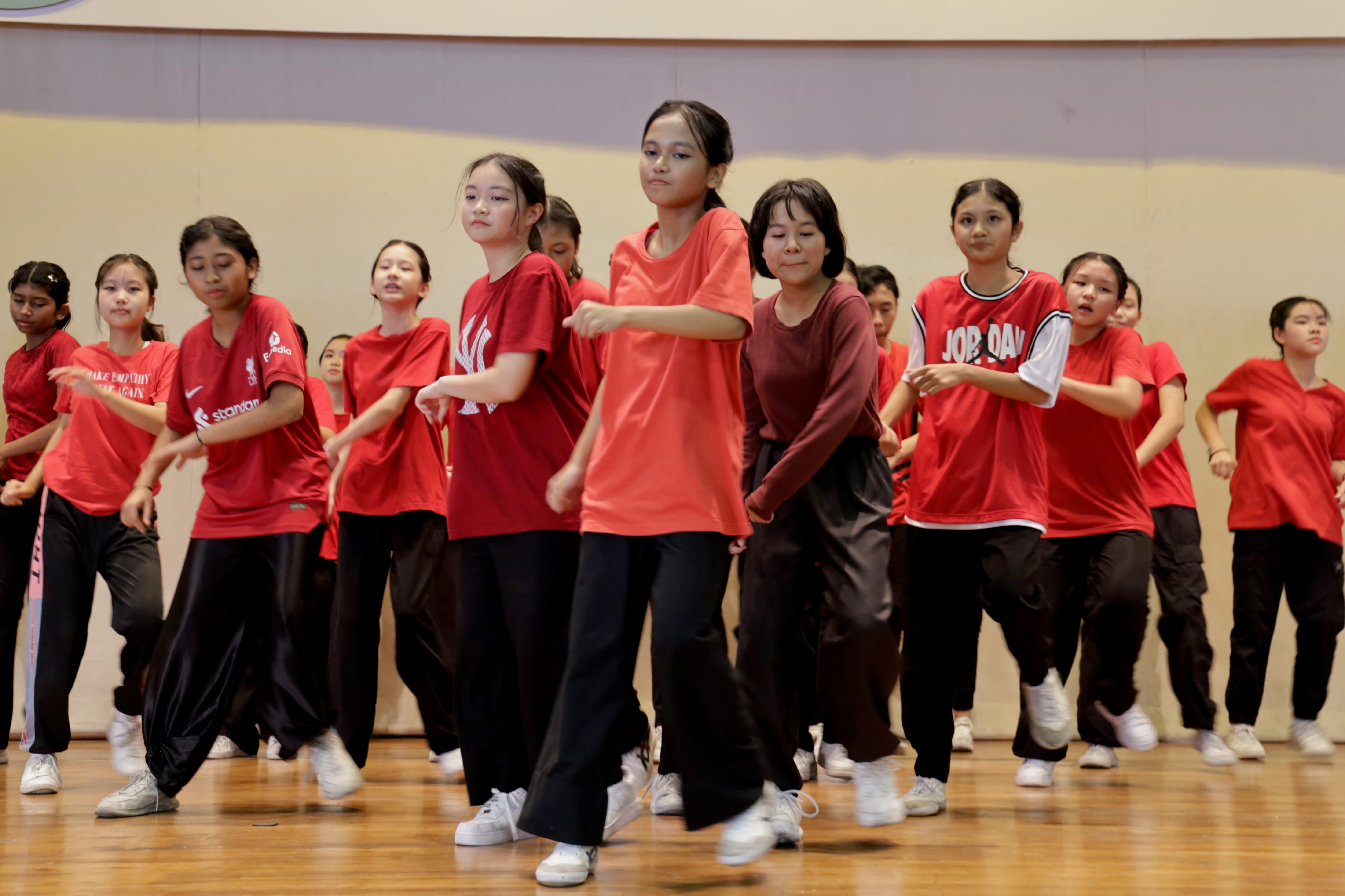 A large group of girls in red shirts and black pants performing a synchronized dance routine on a wooden floor.