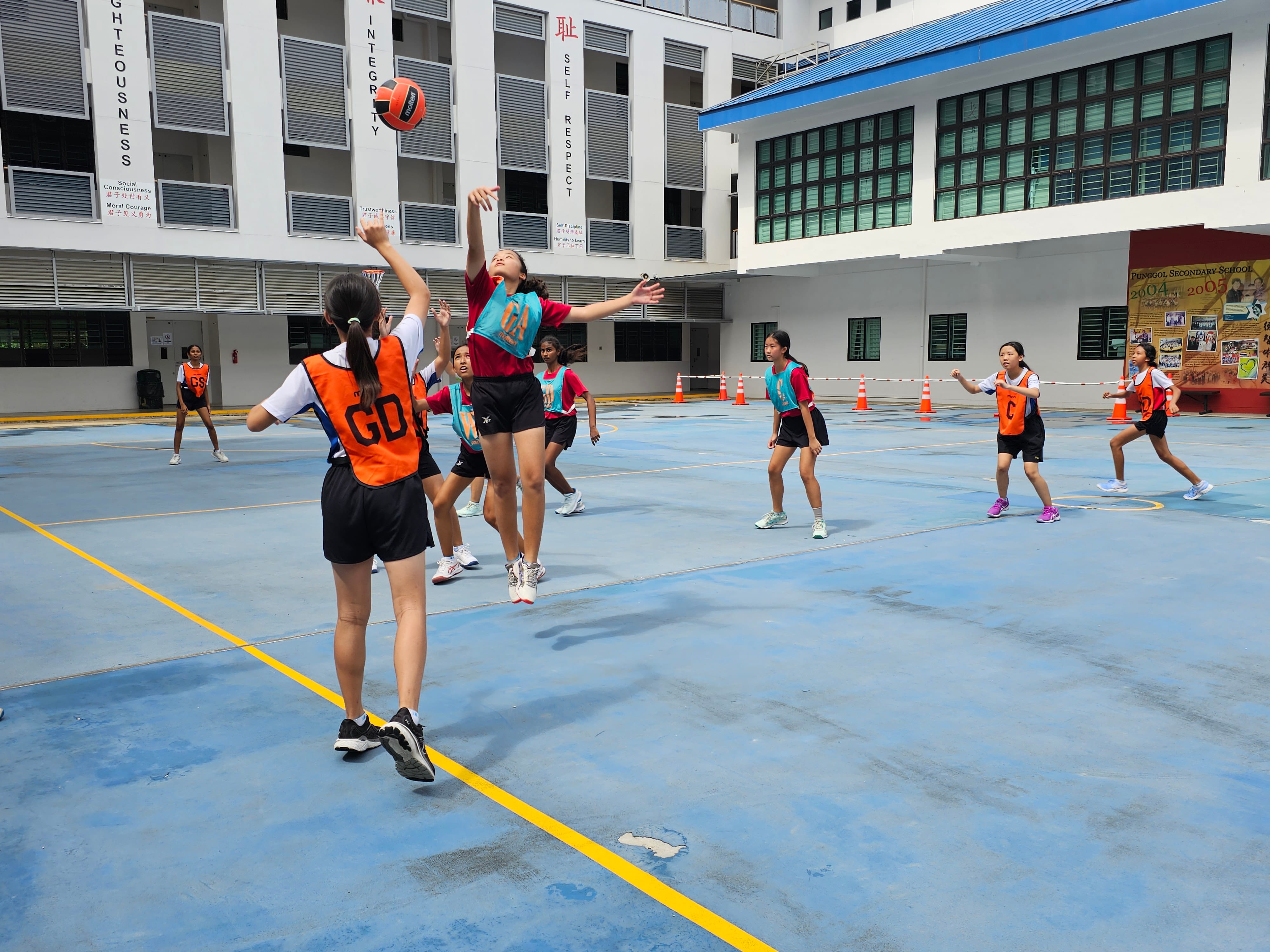 Girls playing netball on an outdoor school court, one player jumping to catch a ball while others in orange bibs defend.