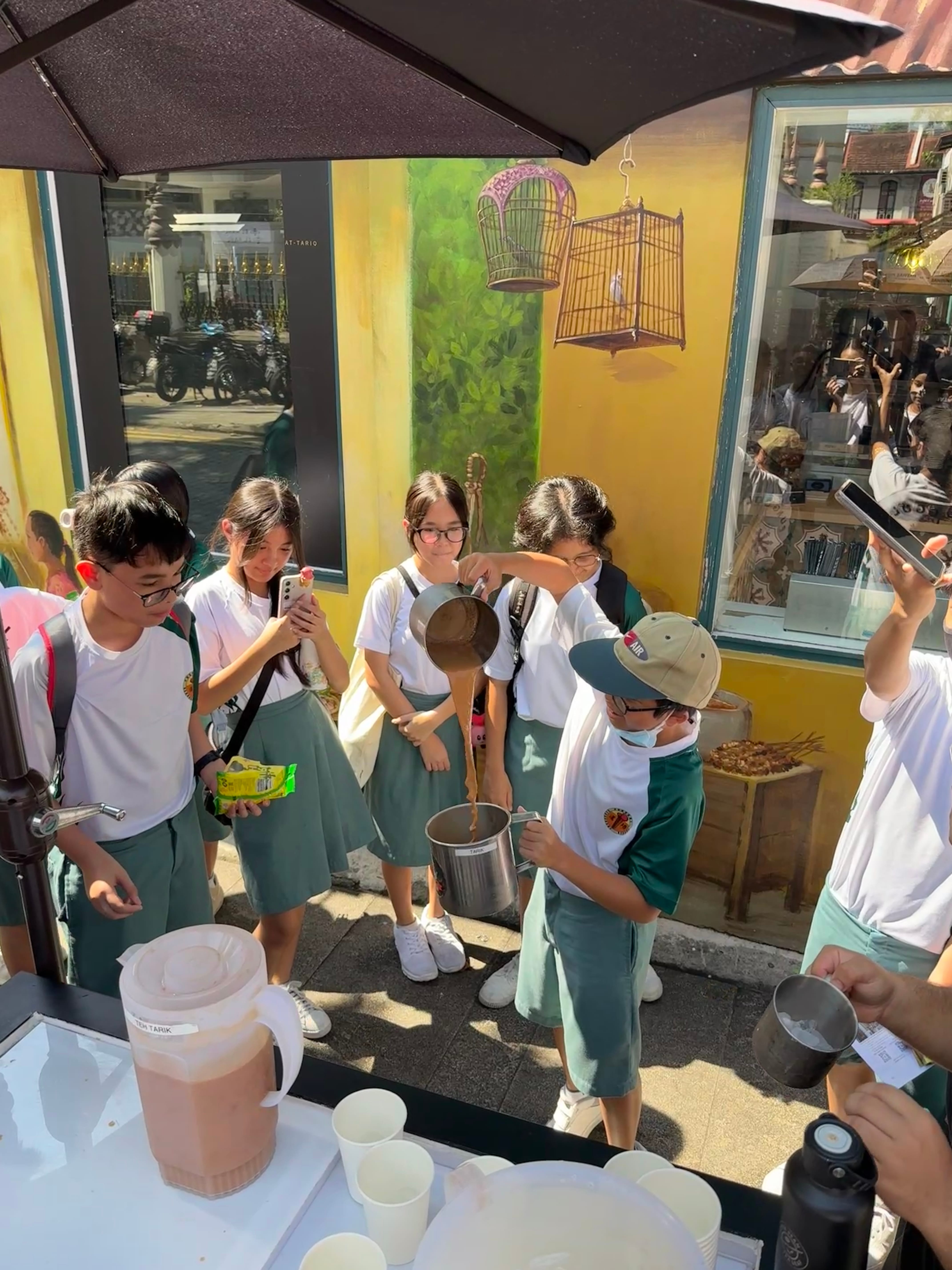 Students in white and green uniforms gather outdoors as one pours a brown liquid between metal containers.