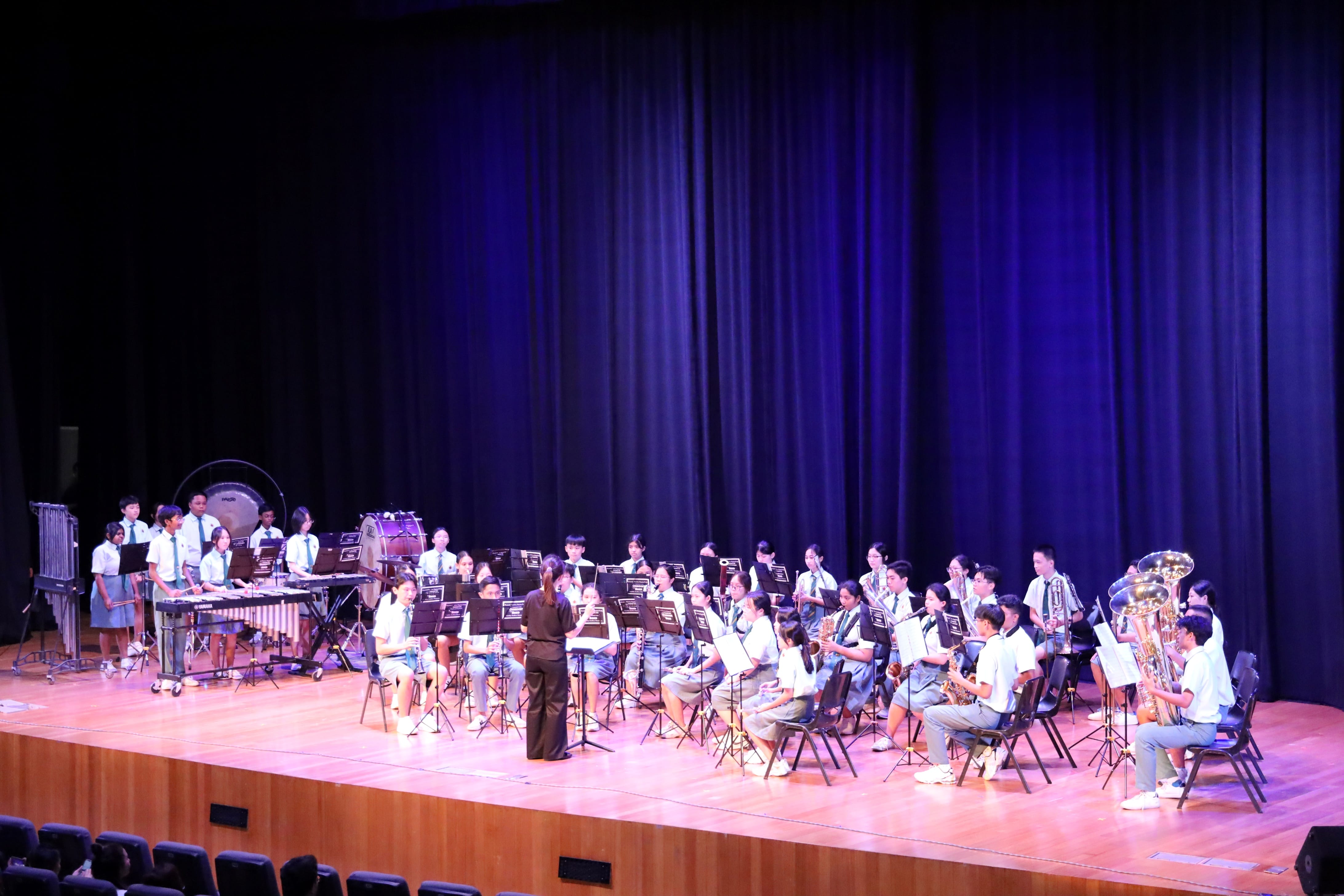 A student band performs on a wooden stage with a conductor, blue curtain backdrop, and various wind and percussion instruments.