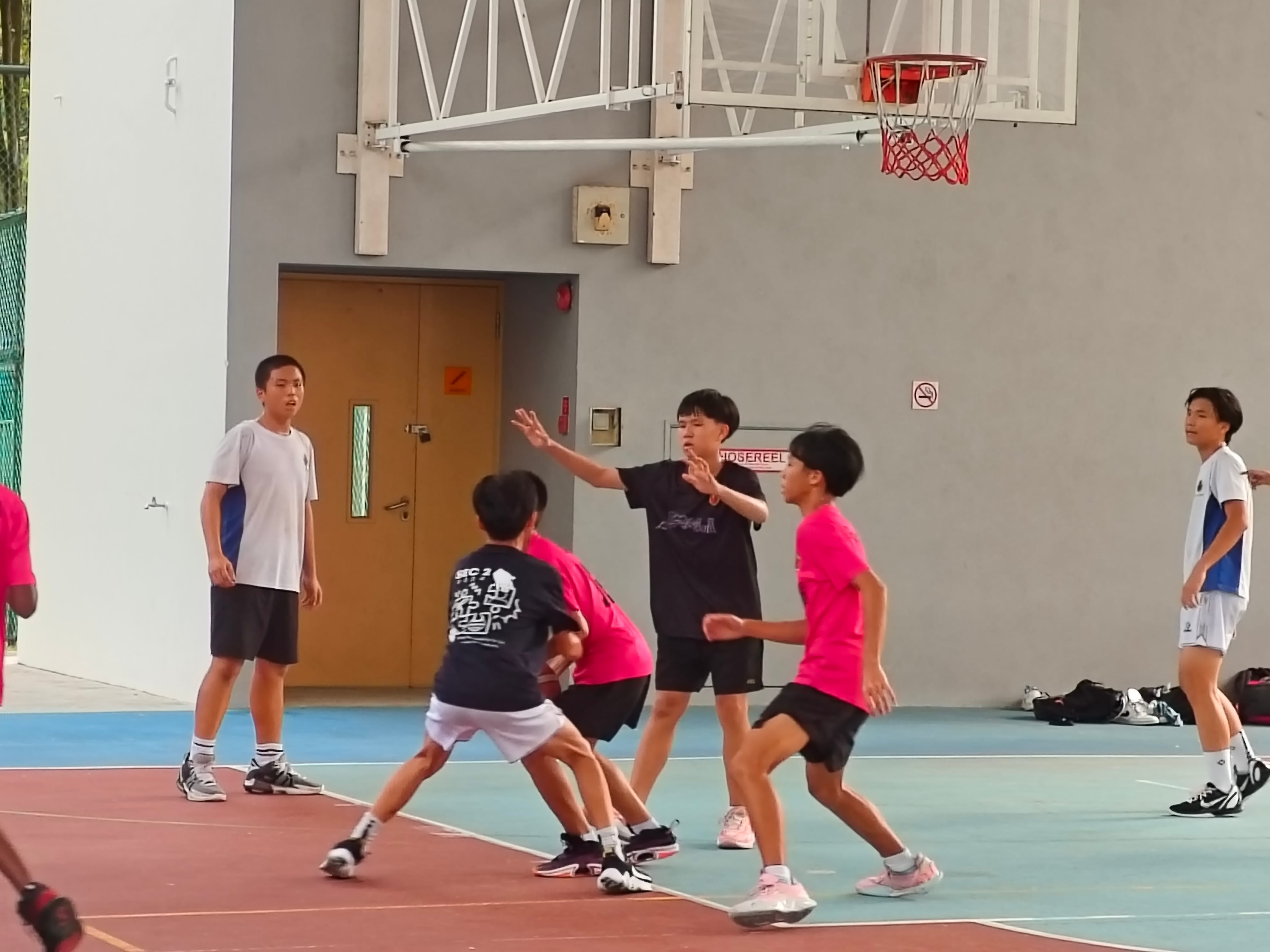 Several young boys playing basketball on an outdoor court with a hoop mounted on a gray wall behind them.