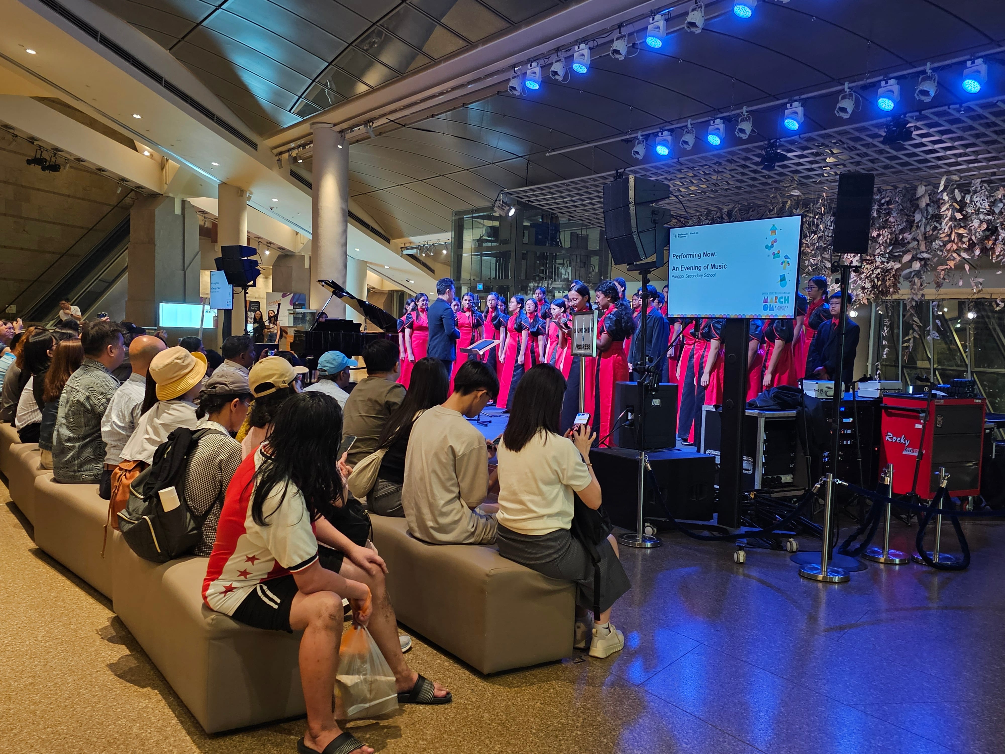 A choir in red and black outfits performs on an indoor stage while an audience sits on benches watching.
