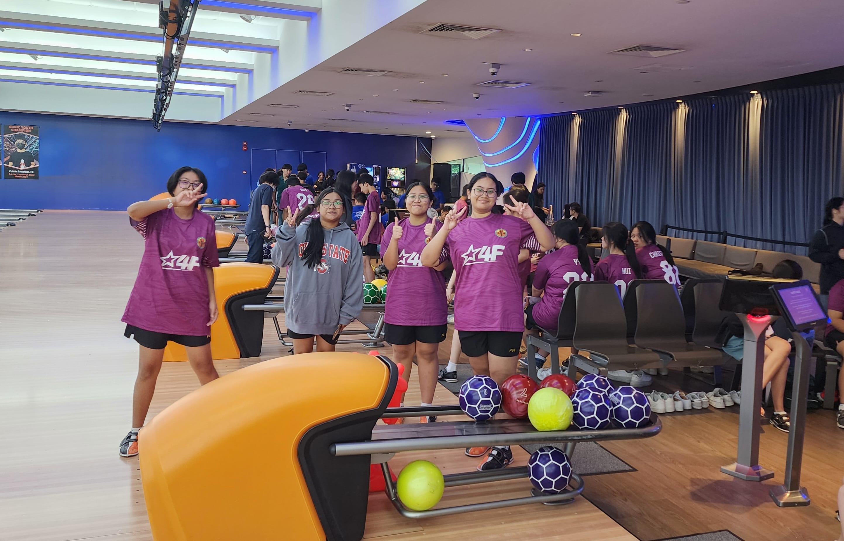 Four teens in purple jerseys pose and smile near a bowling ball rack inside a busy bowling alley.