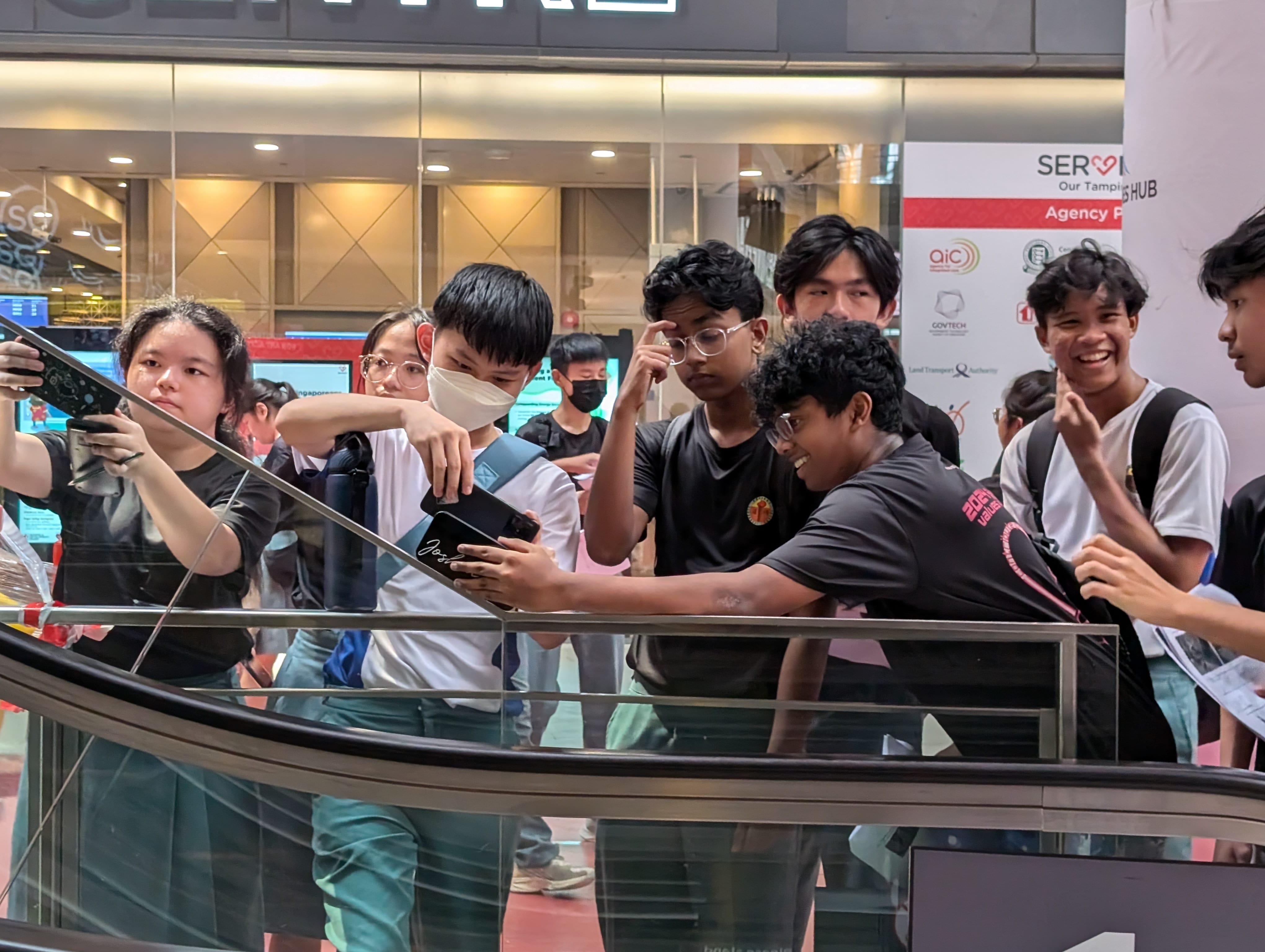 A group of teenagers gathered around a railing inside a mall, looking at a phone, with a banner behind them.