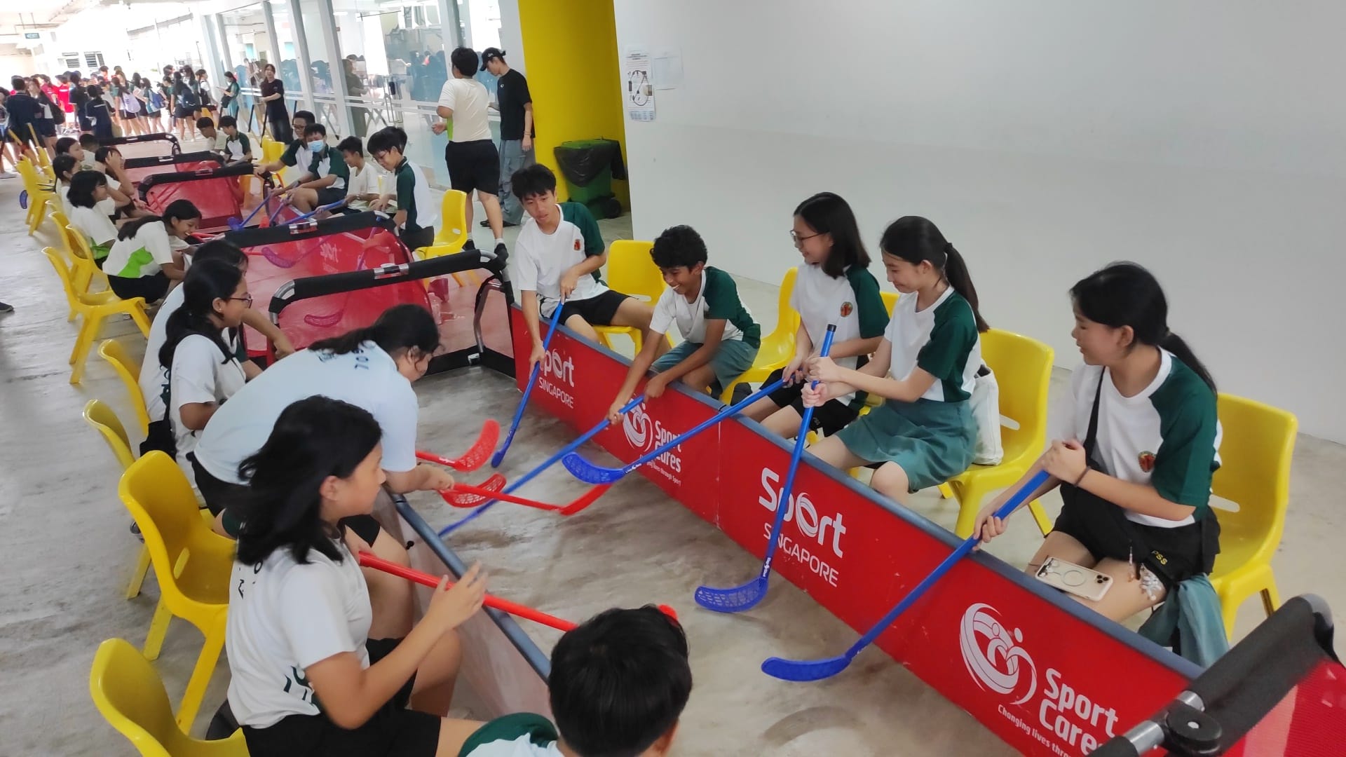 Students sit on yellow chairs playing a seated floorball activity at a Sport Singapore SportCares event indoors.