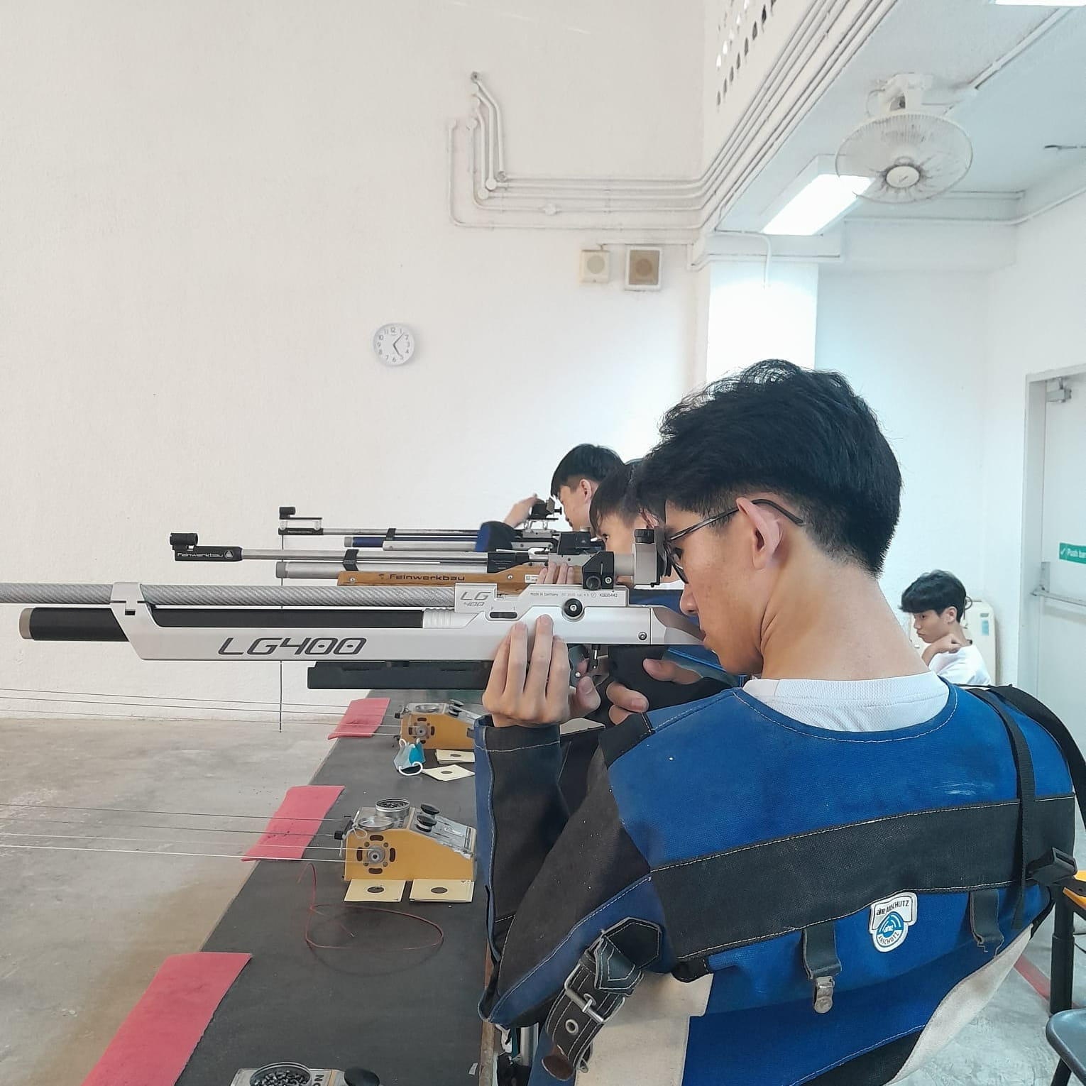 Three young men in shooting vests aim Feinwerkbau LG400 air rifles at an indoor shooting range.