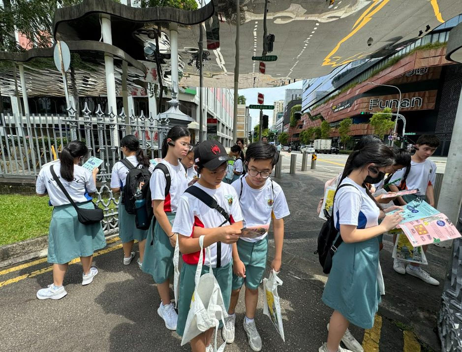 Students in white uniforms and teal skirts or shorts stand on a Singapore street near the Funan mall, reading maps.
