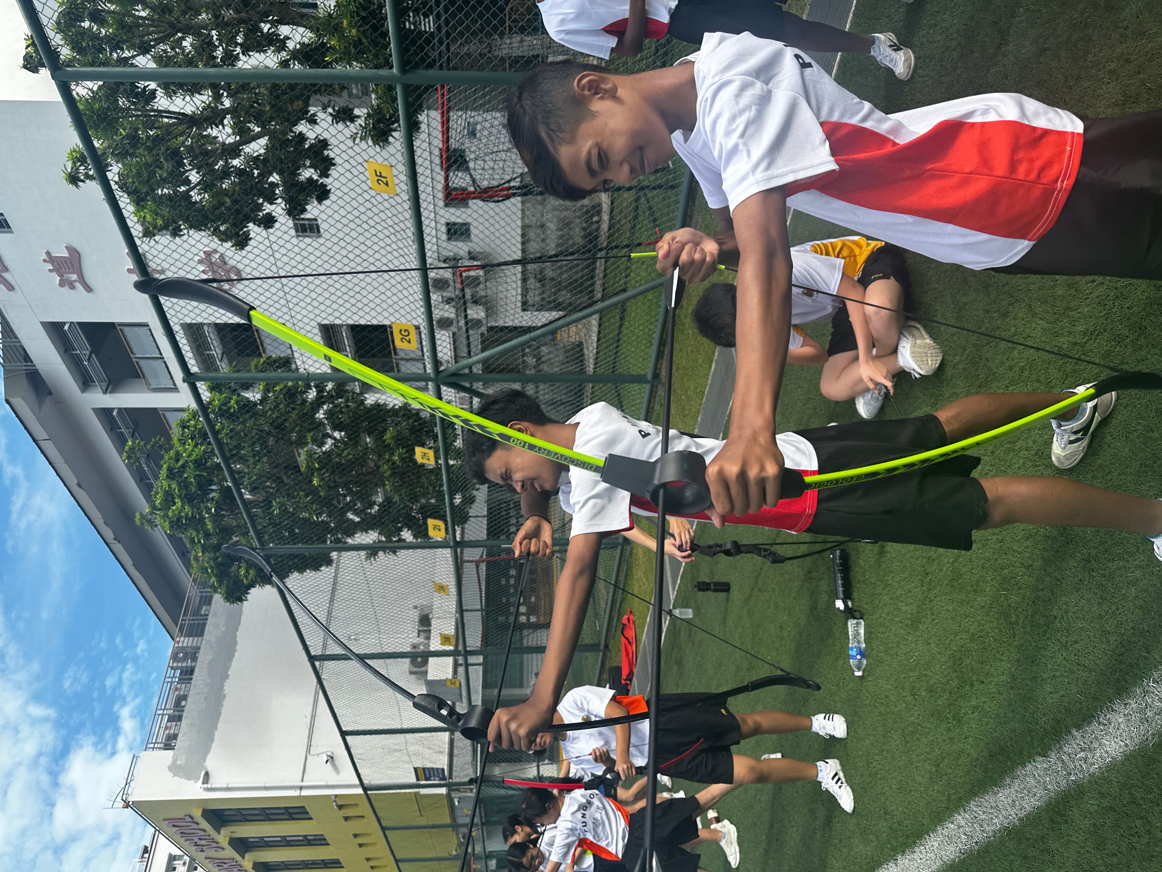 Students in white uniforms practice archery with yellow bows on a school sports field outdoors.