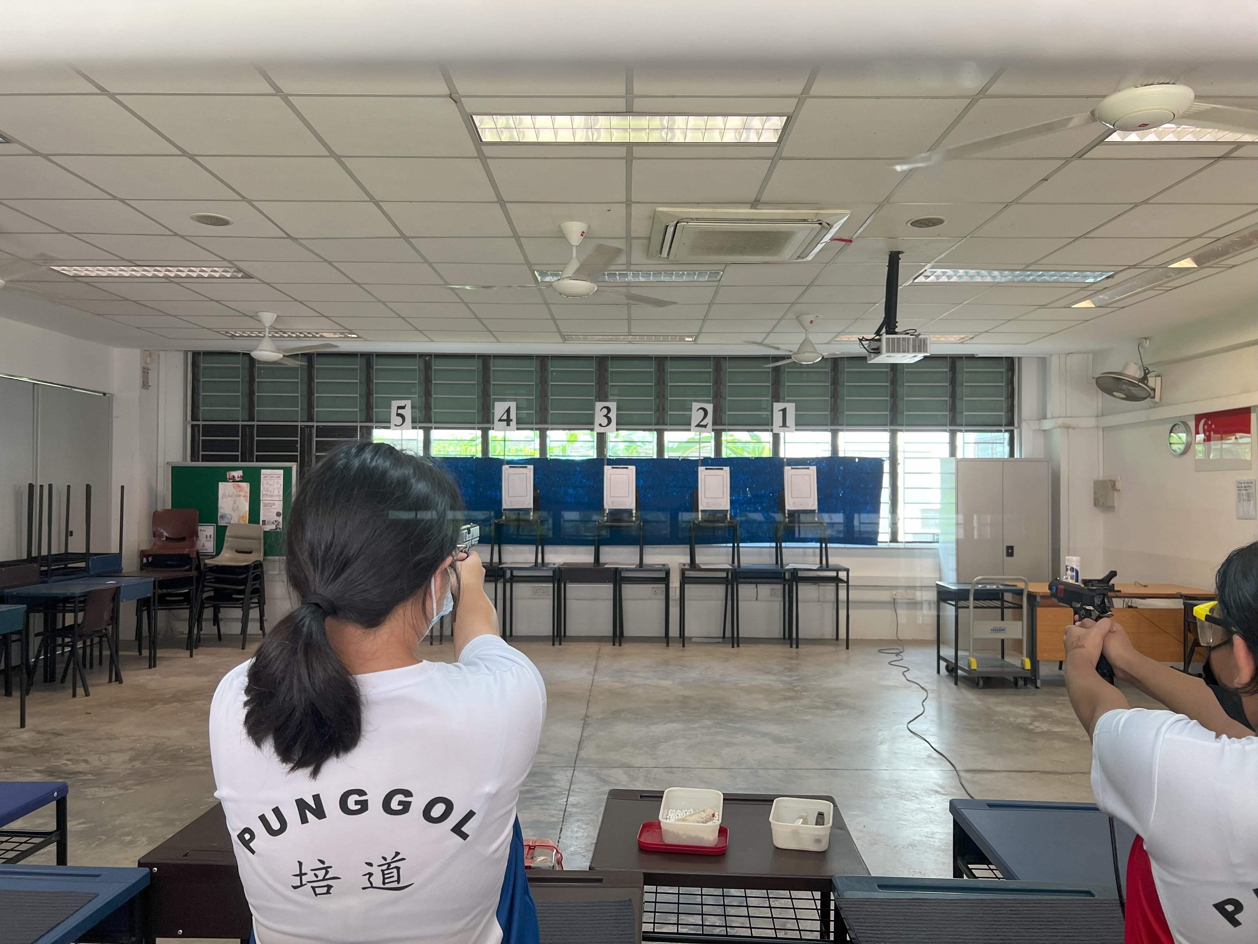Two people in Punggol shirts aim pistols at numbered paper targets on an indoor shooting range.