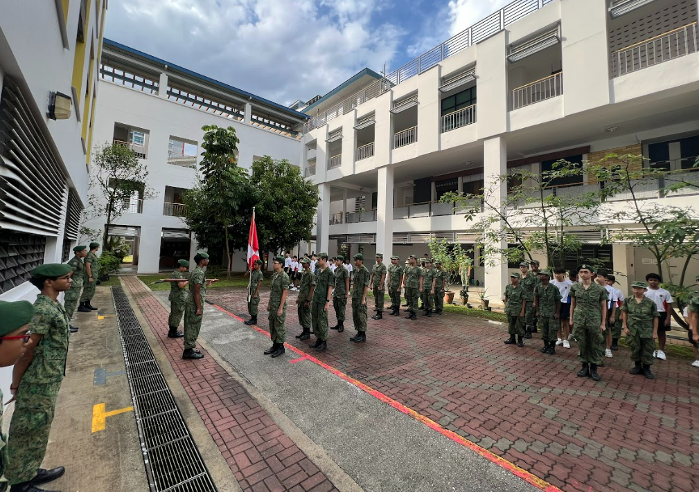 Soldiers in green camouflage uniforms stand in formation in a school courtyard, with a red and white flag bearer.