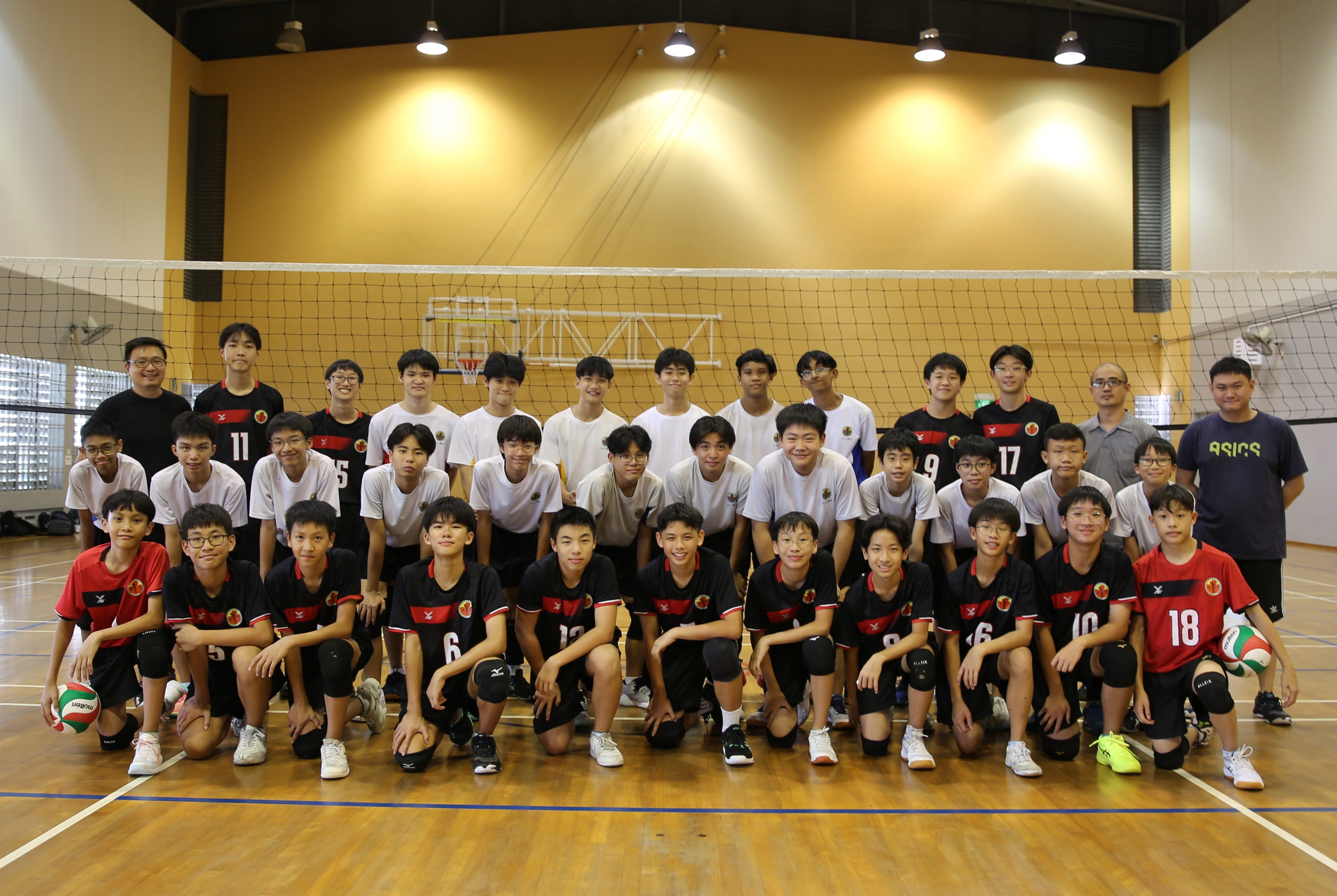 A youth volleyball team in black and red uniforms poses for a group photo indoors in front of a net.