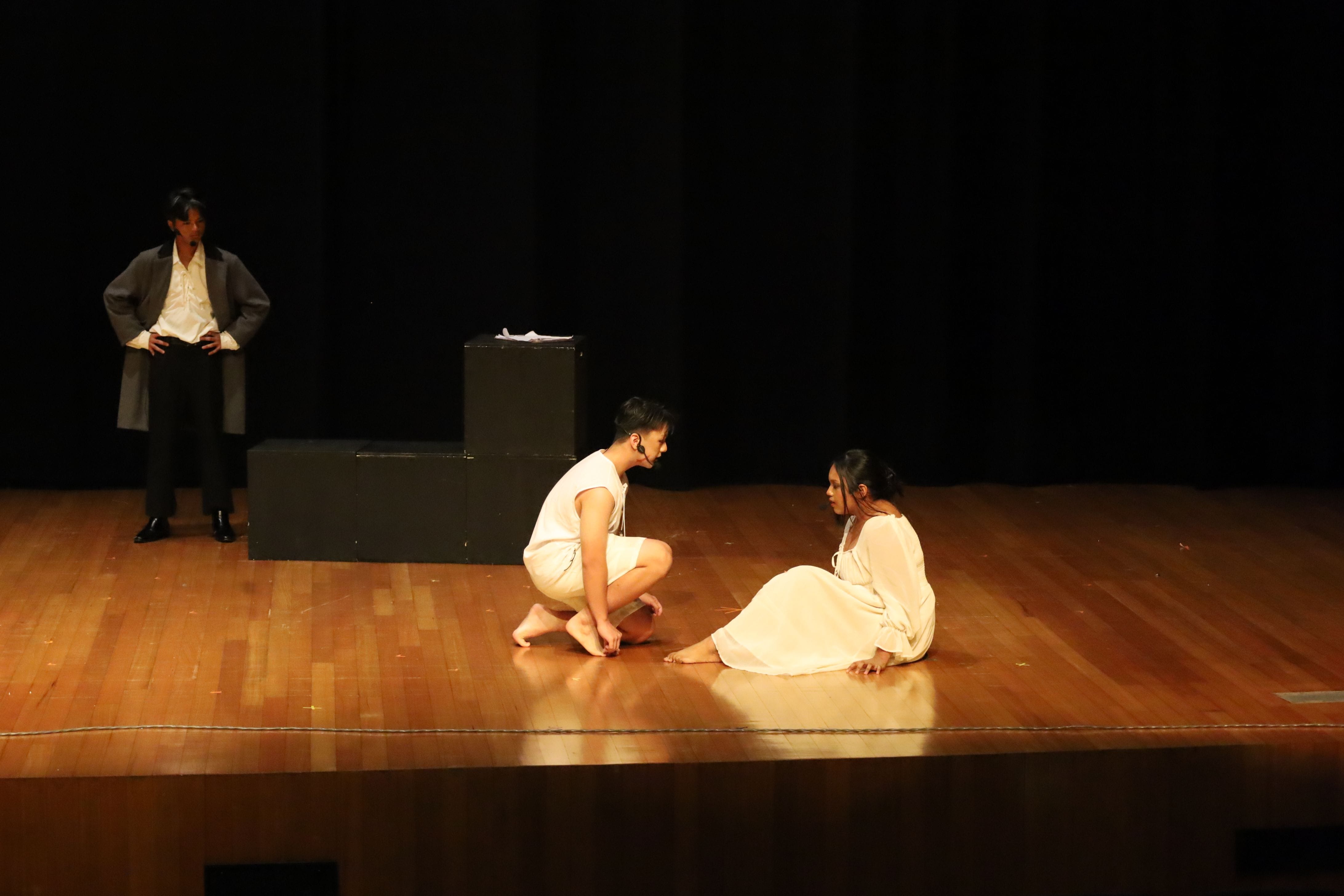 Three performers on a dark stage; two in white sit on the floor facing each other while one in gray stands watching.