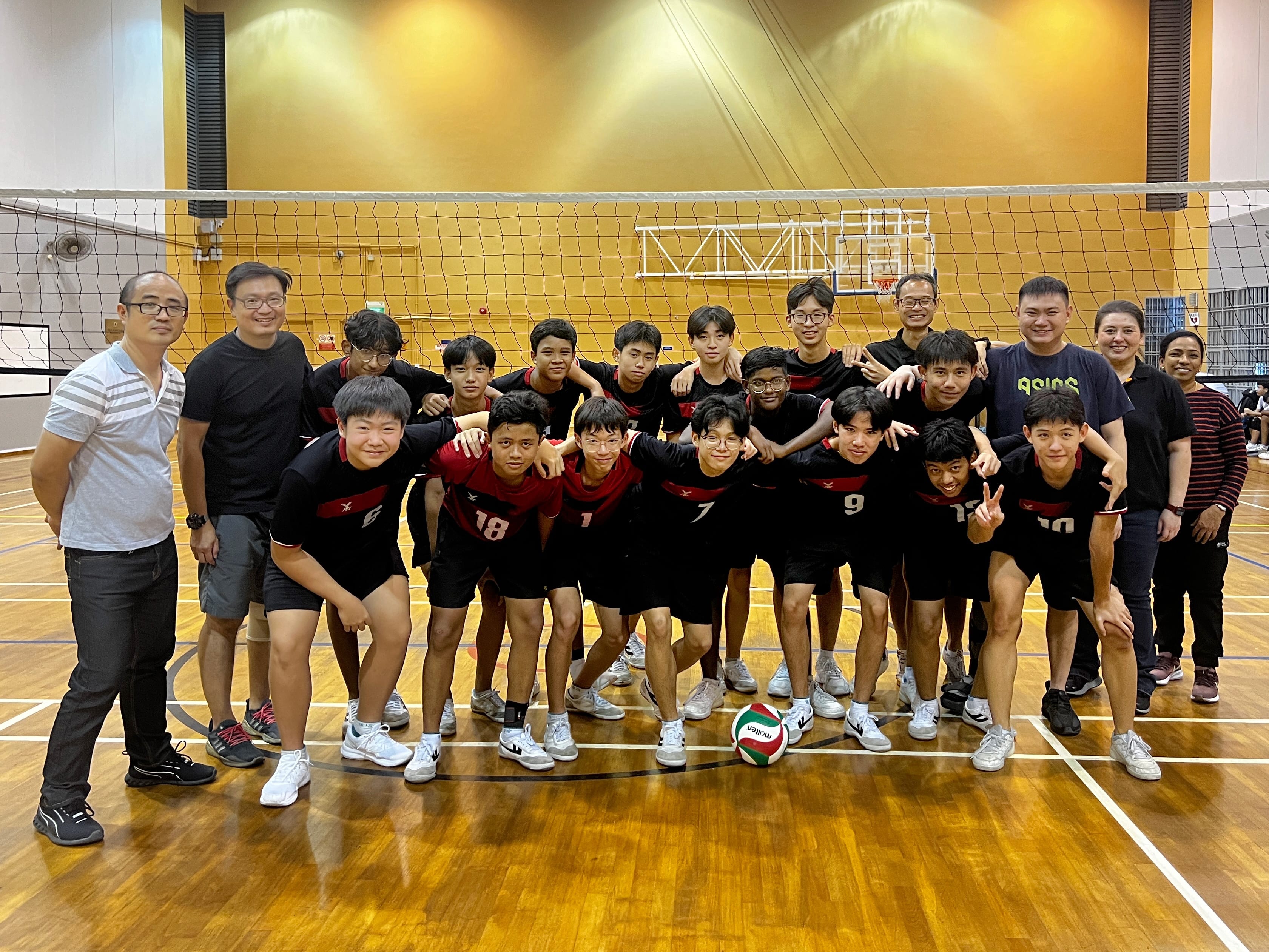 A youth volleyball team in black and red jerseys poses with coaches in an indoor gym near a net with a ball on the floor.
