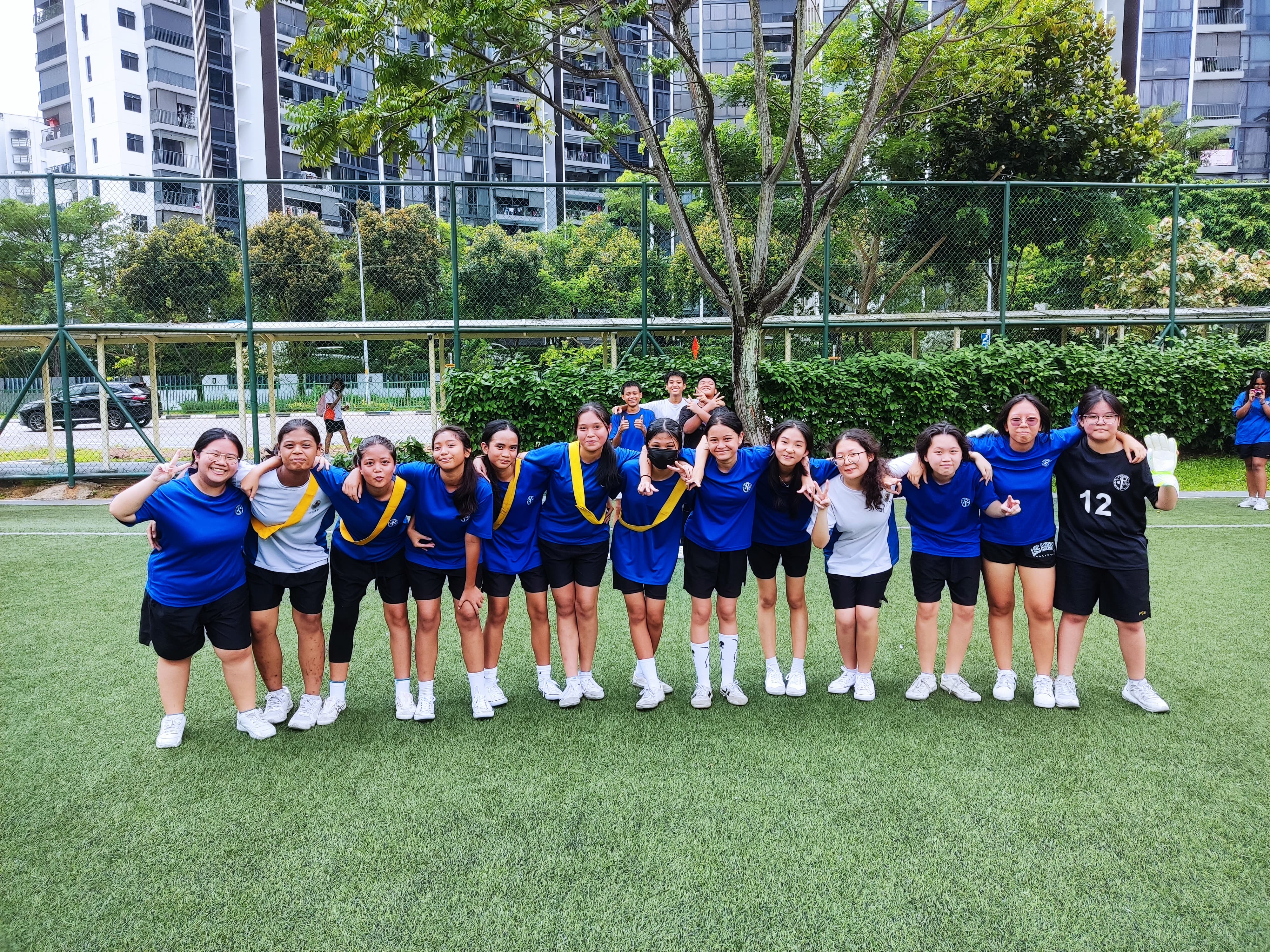 A group of young girls in blue sports uniforms posing together on a green turf field outdoors.