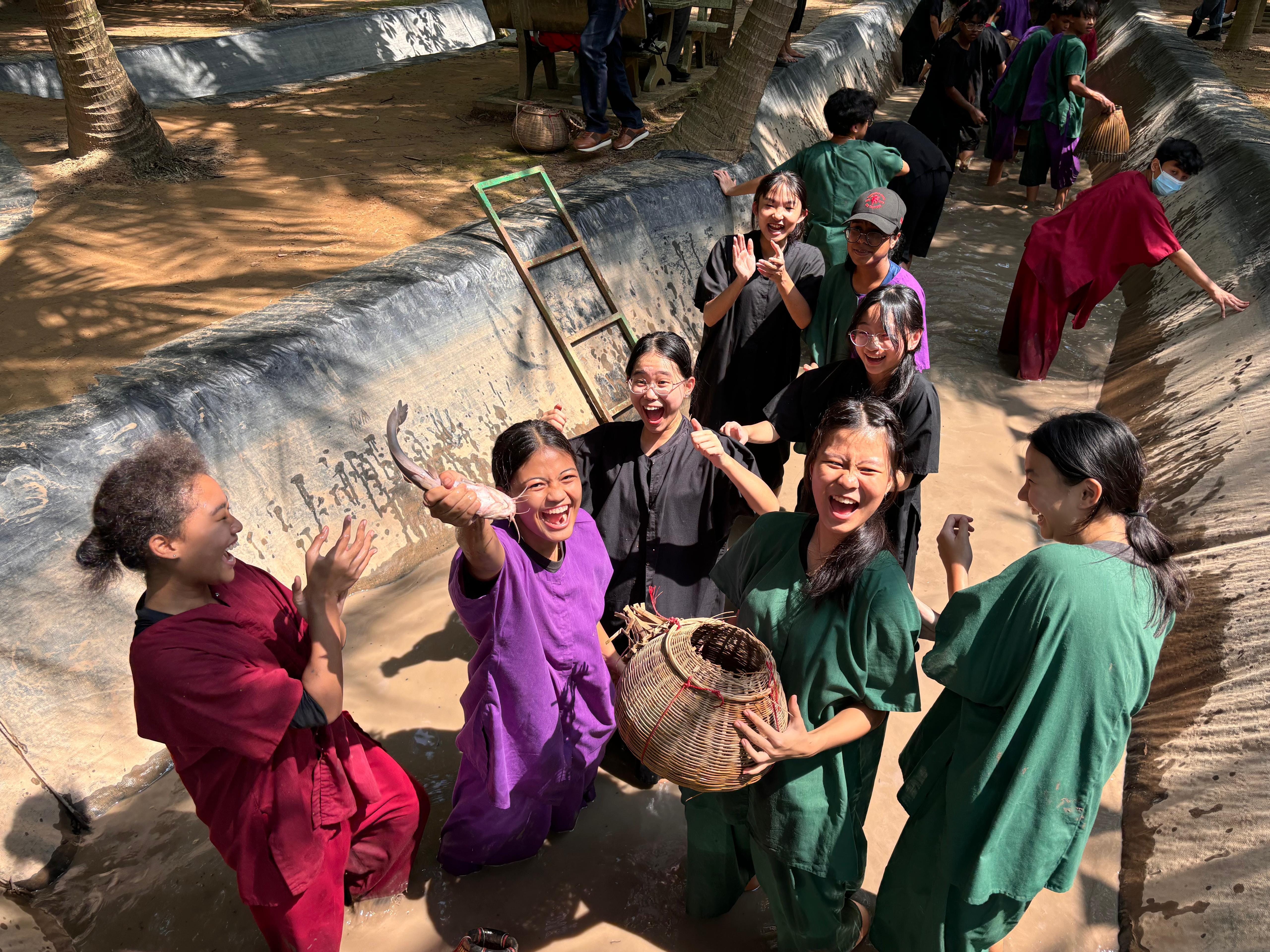 A group of laughing young people in colorful traditional clothing hold a fish and wicker basket outdoors near a canal.
