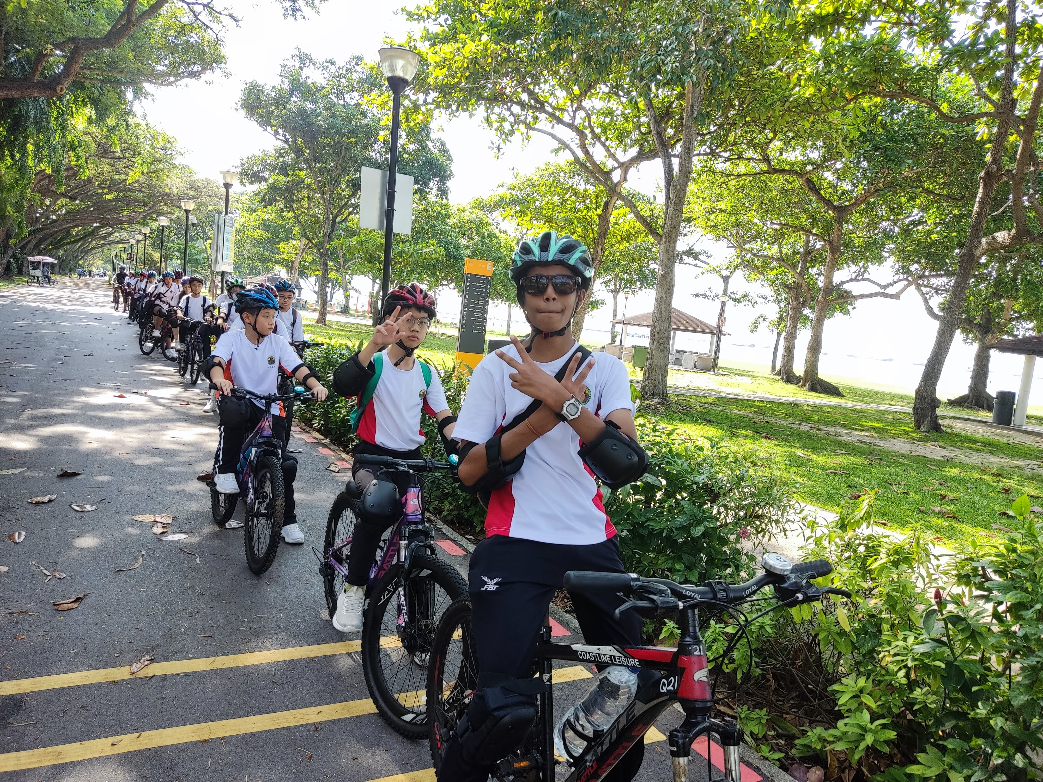 A group of students in helmets pose with bicycles on a tree-lined park path near a waterfront.