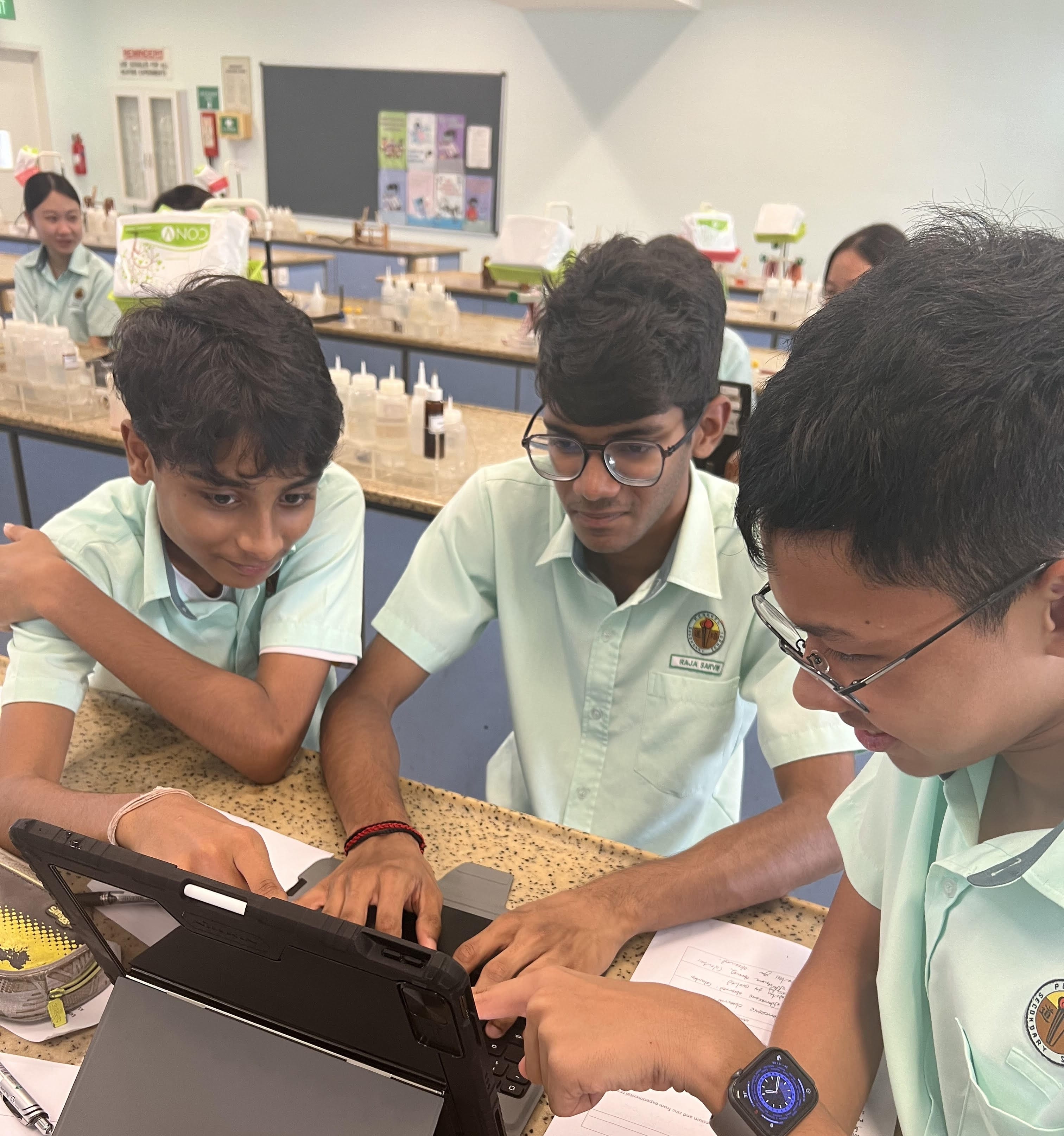 Three students in light blue uniforms huddle around a tablet on a lab bench in a science classroom.