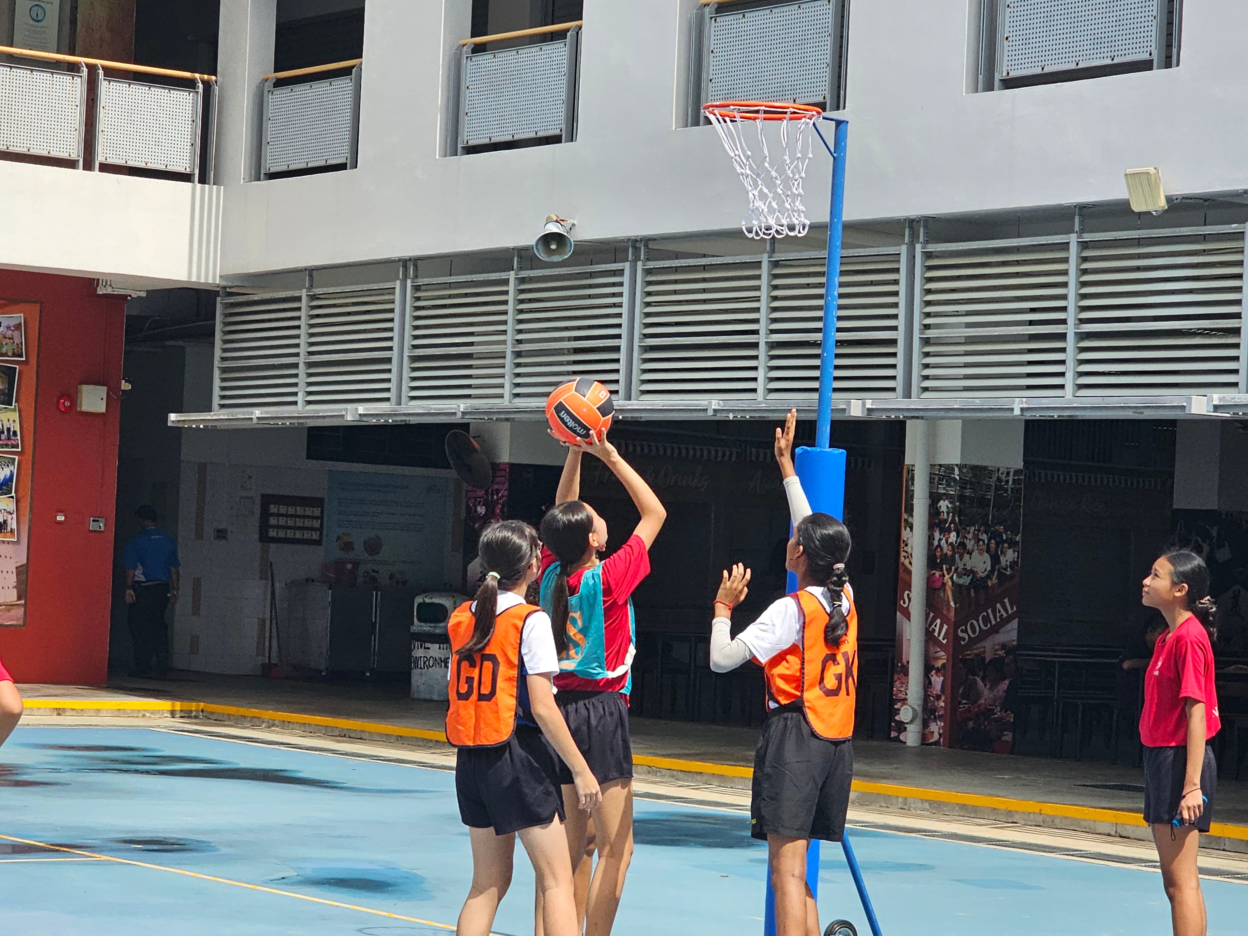 Girls playing netball on an outdoor court, one shooting toward the hoop while defenders in orange vests try to block.