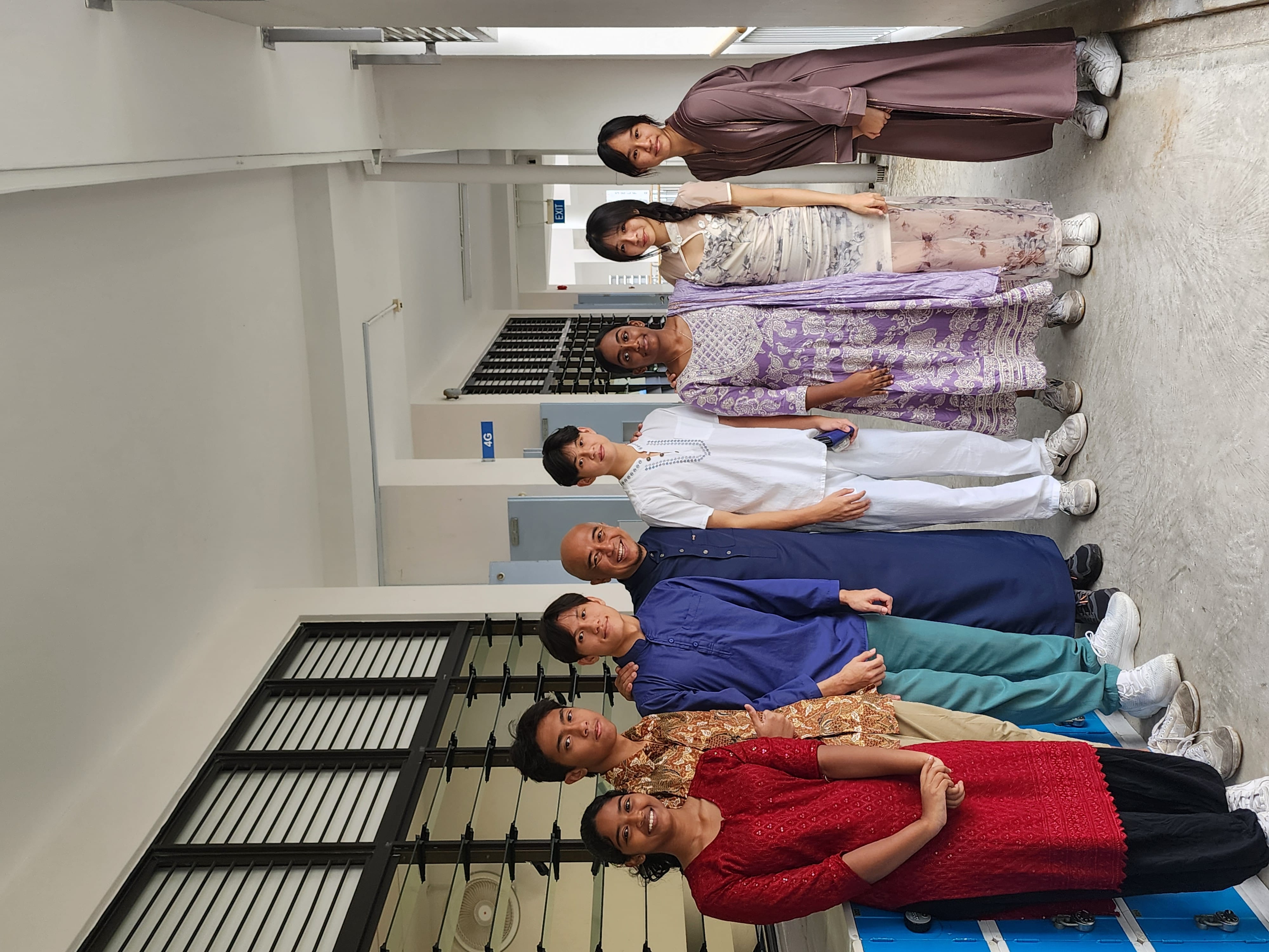 Eight people in colorful traditional outfits pose together smiling in a school corridor with lockers.