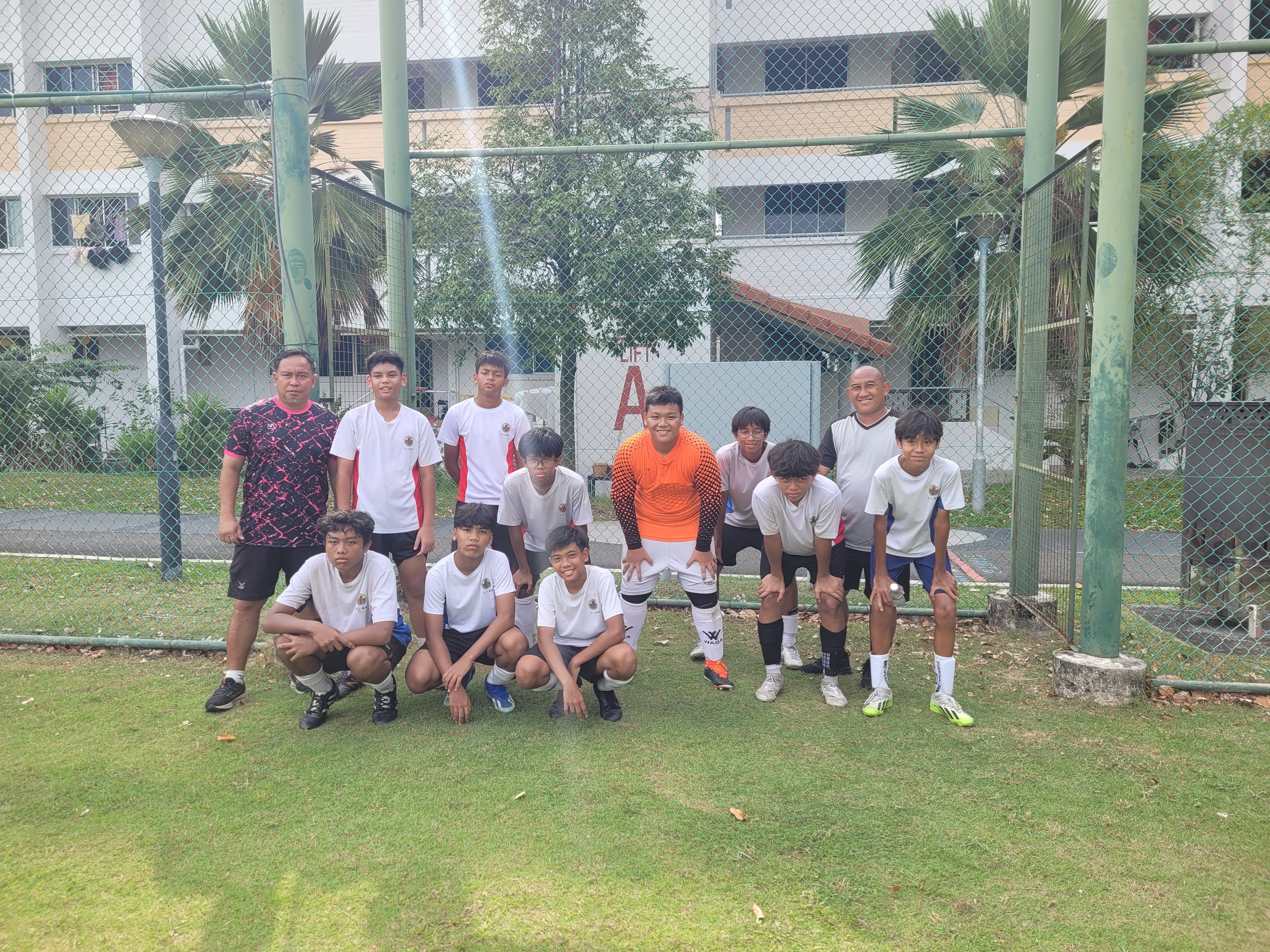 A youth football team in white jerseys poses on grass near a chain-link fence, with two adult coaches on either side.