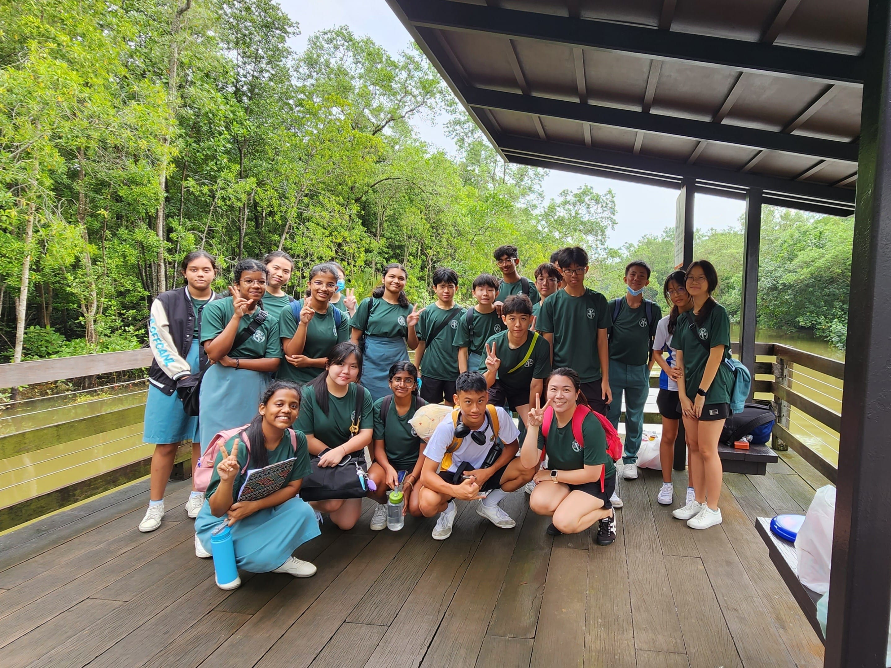 A group of students in green uniforms pose on a wooden deck shelter surrounded by lush green trees.