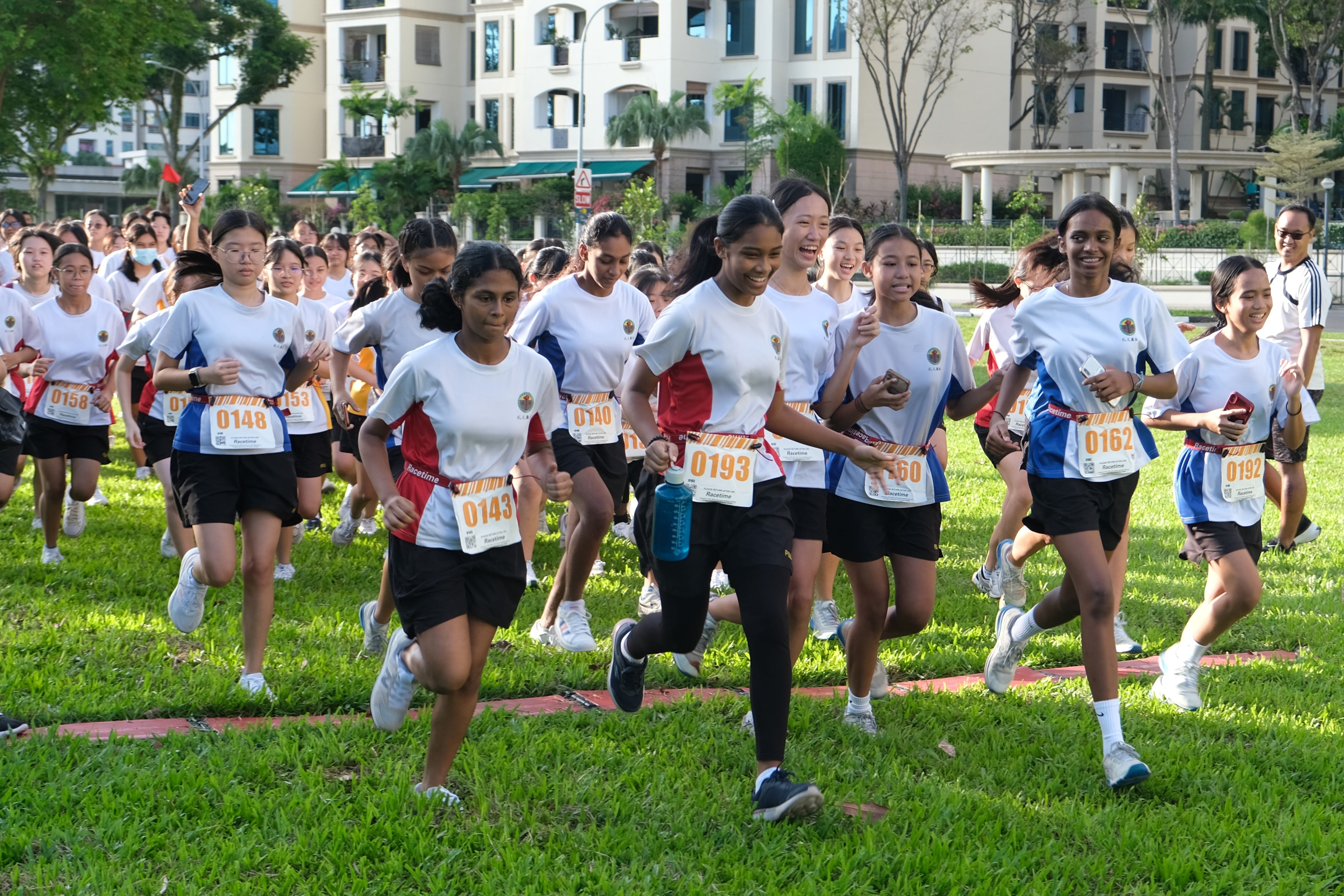 A large group of girls in white uniforms with numbered race bibs running on grass during an outdoor race event.