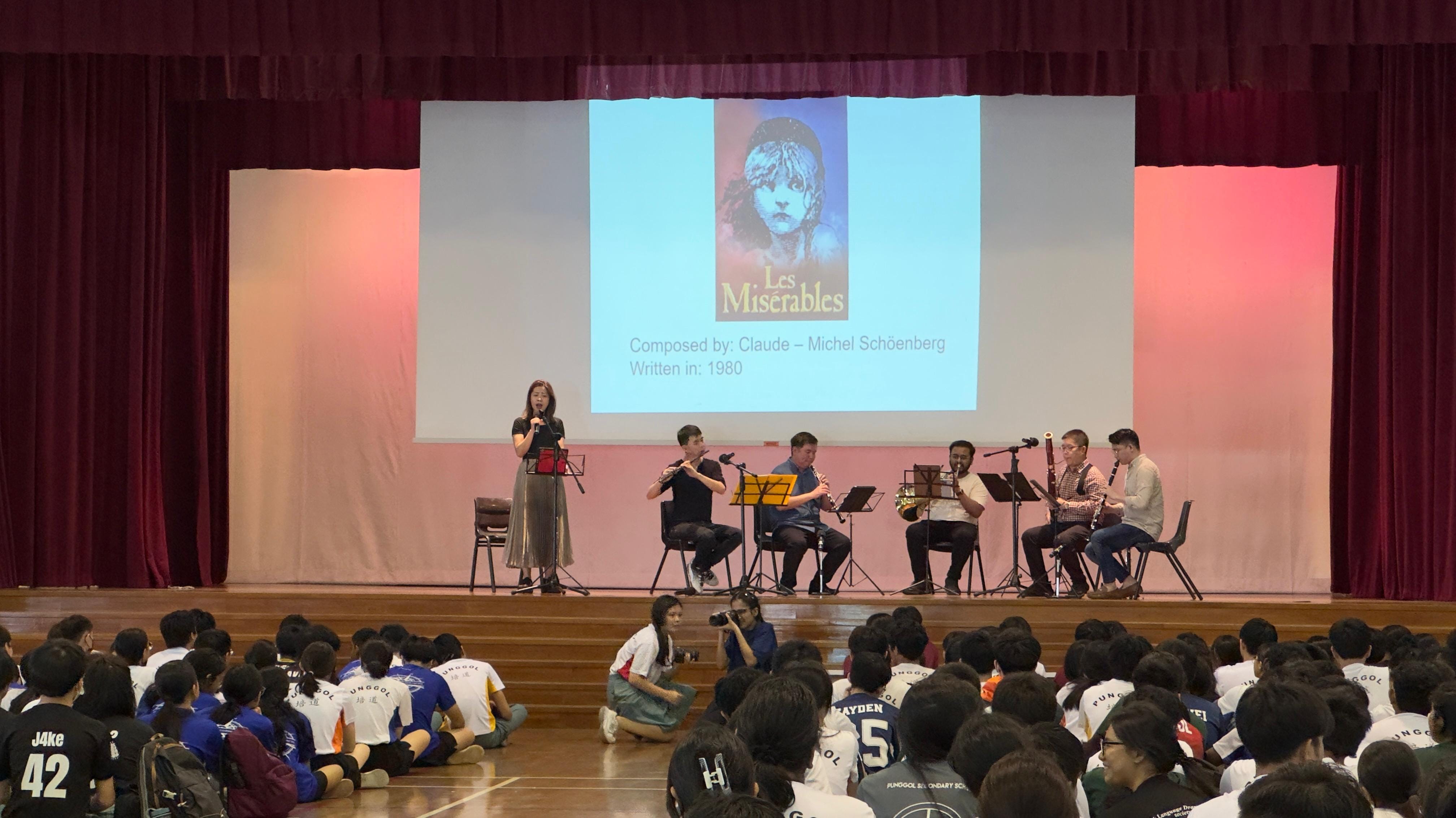 A woman sings on stage with musicians as students sit on the floor watching, with a Les Miserables slide projected behind.