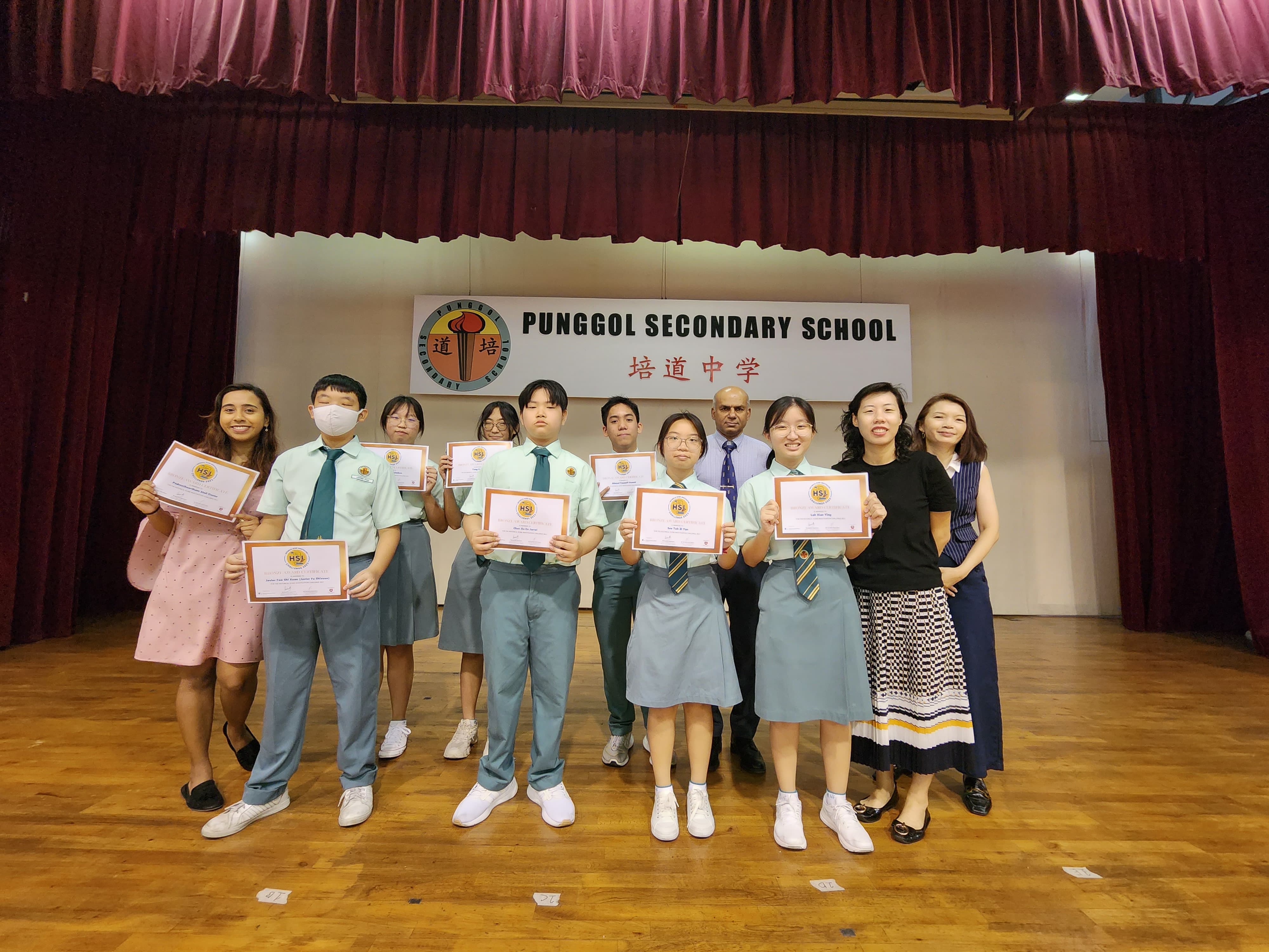 Students and teachers pose on a stage holding certificates in front of a Punggol Secondary School banner.