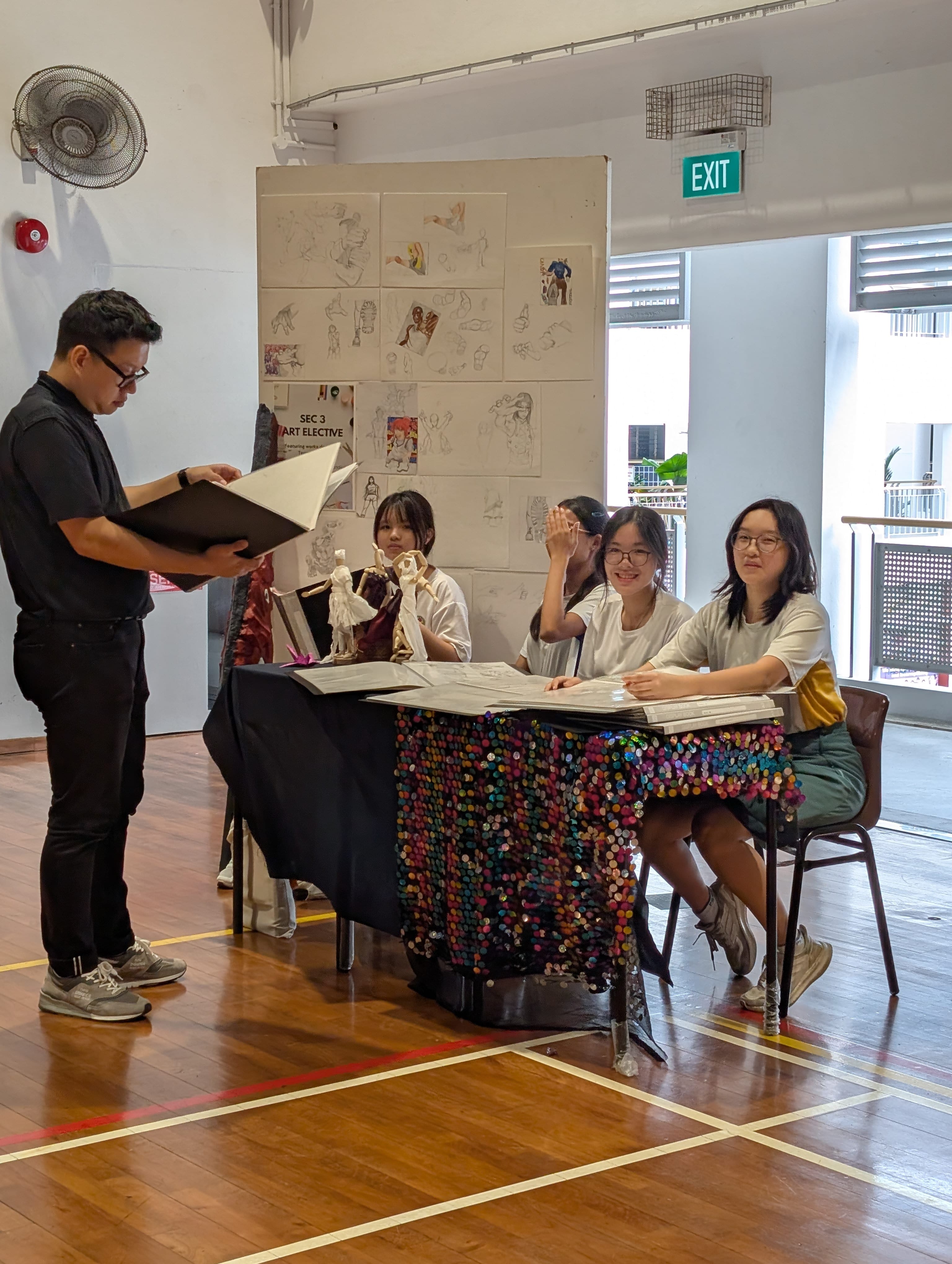 A man reviews a portfolio while three students sit at a sequined table displaying art sketches in a school hall.