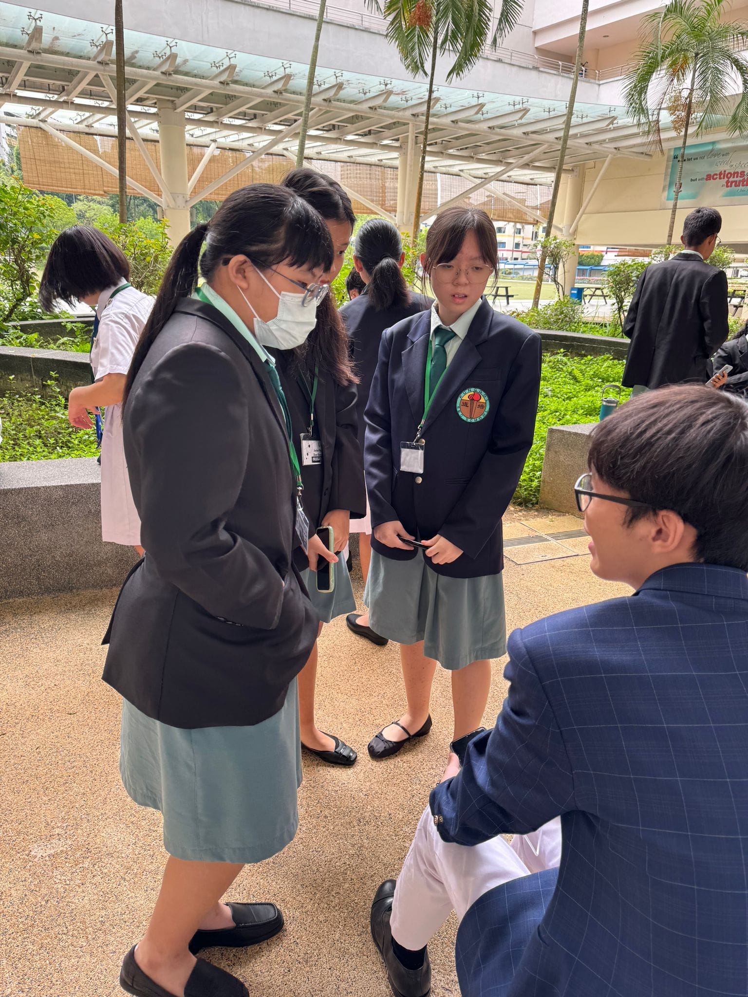 Several school students in navy blazers and green ties stand talking outdoors under a covered walkway.