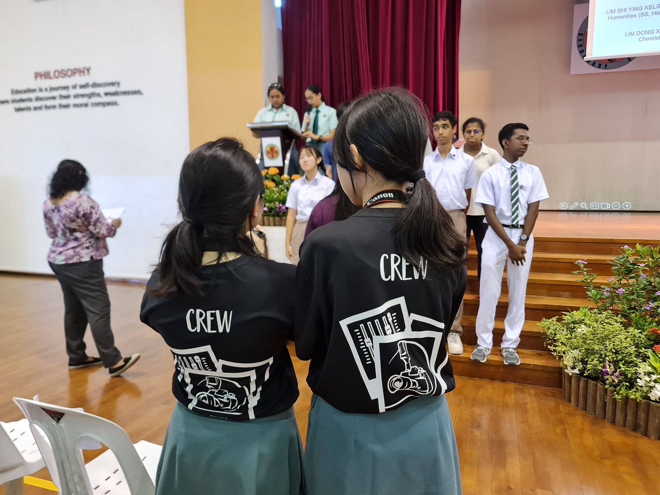 Two people wearing black CREW shirts stand facing a school stage during a ceremony, with students and a podium visible.