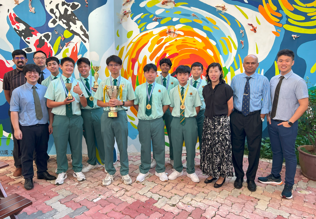 Students in green uniforms with medals and trophies pose with teachers before a colorful mural.