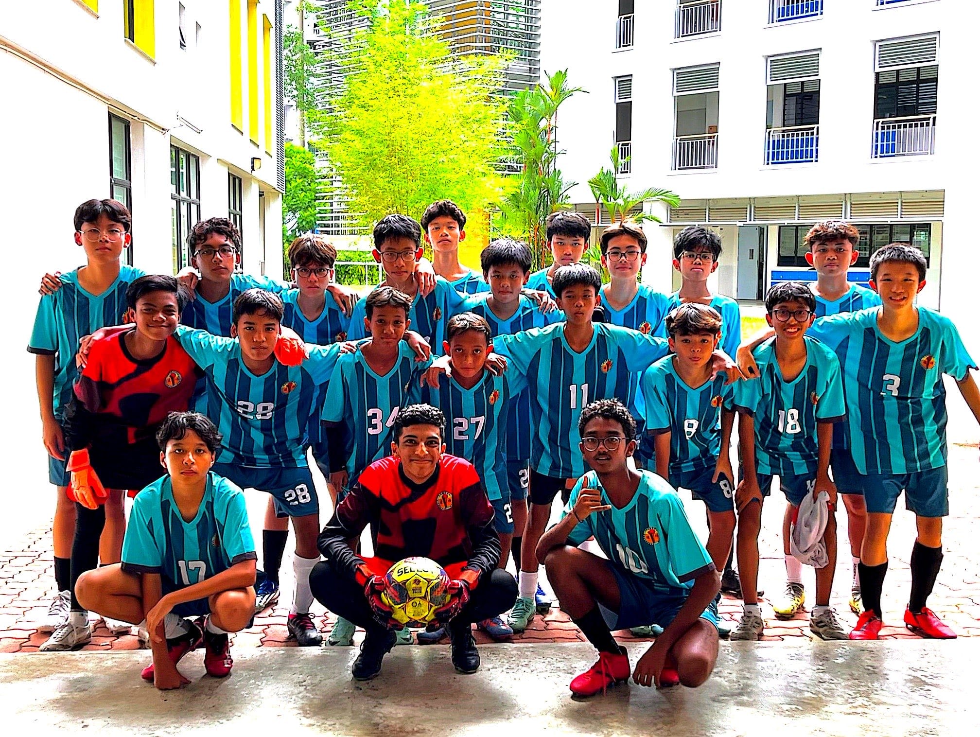 A youth football team of about 18 boys in teal jerseys pose together outdoors, with one holding a soccer ball.