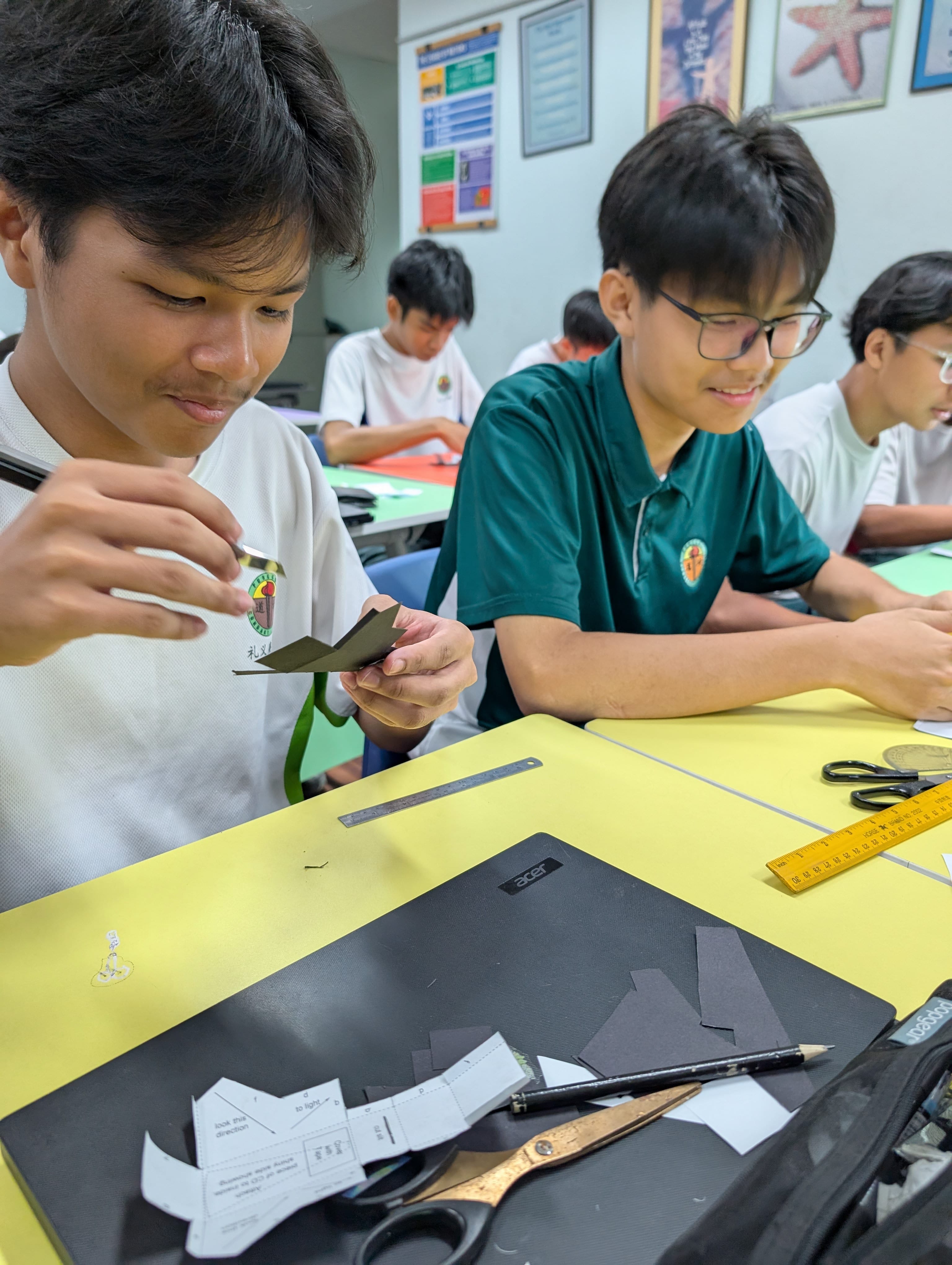 Two male students in uniforms cut and fold dark paper at a yellow desk with scissors, a ruler, and a laptop.