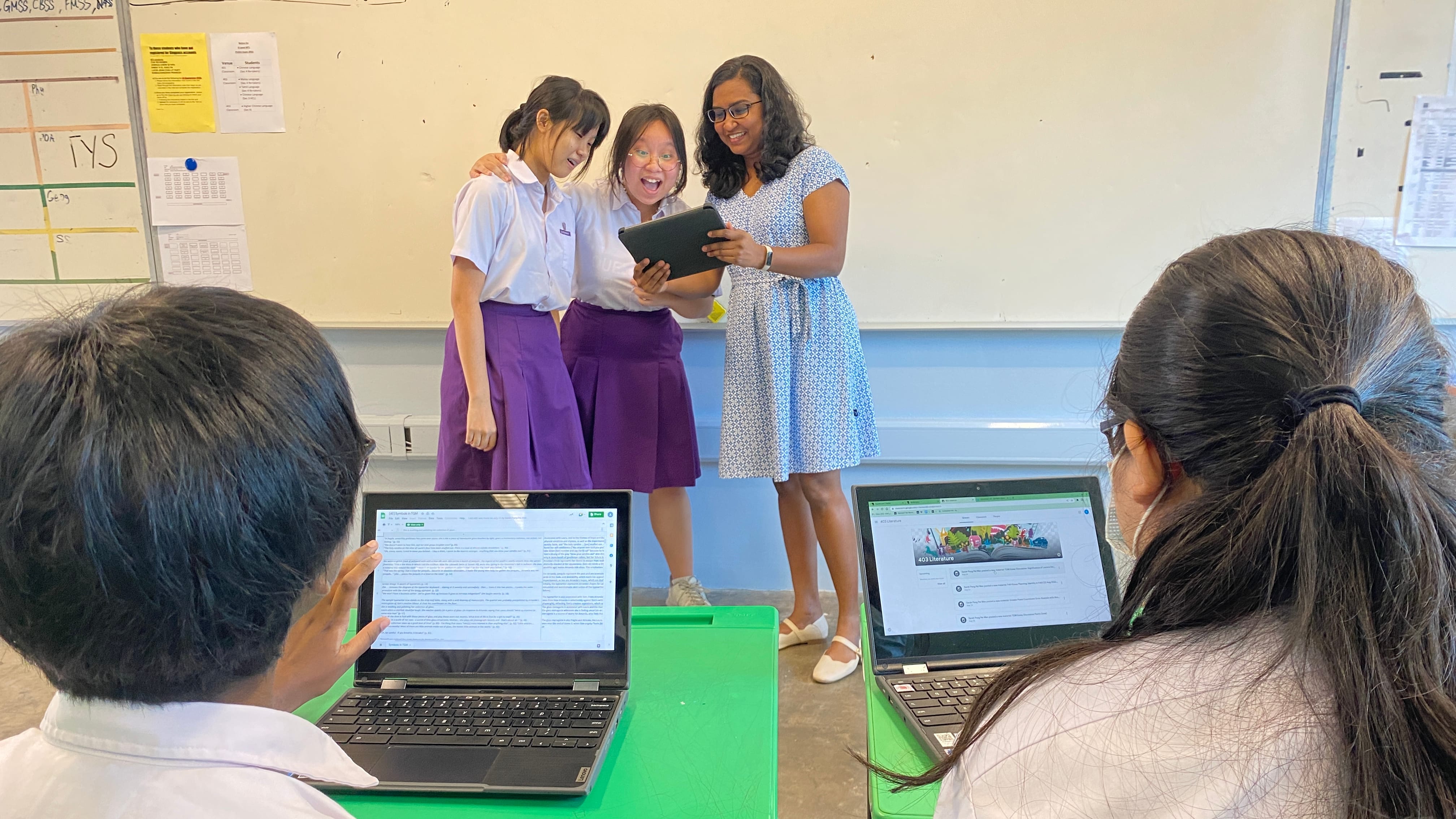 A teacher and two uniformed students look at a tablet in a classroom while two other students use laptops nearby.