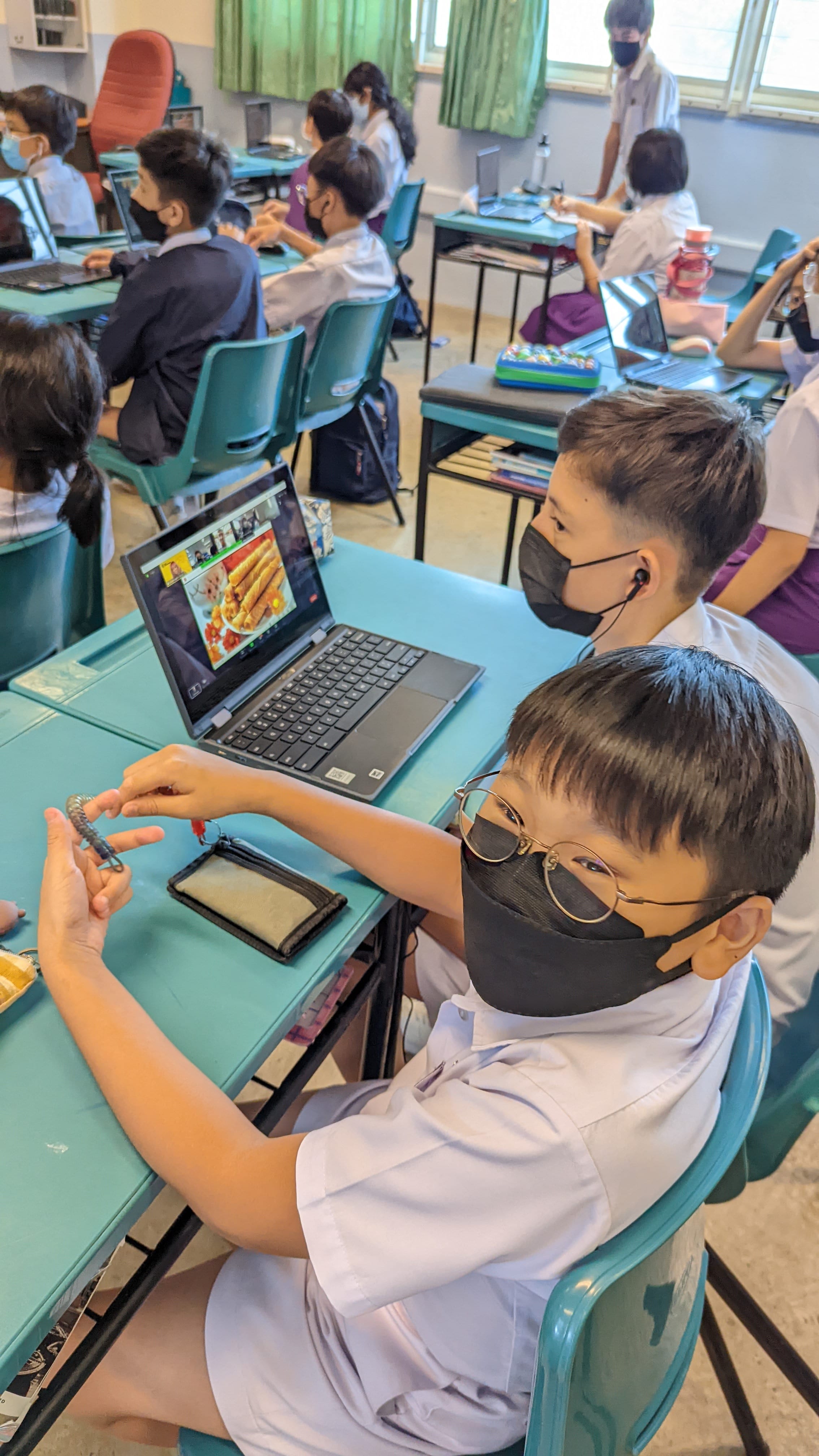 Students in masks sit at teal desks in a classroom, one boy with glasses holds a keychain near an open laptop.