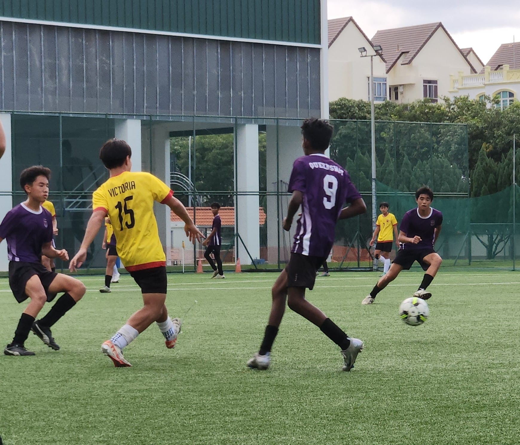 Boys in yellow and purple jerseys compete for a soccer ball on a green artificial turf field.