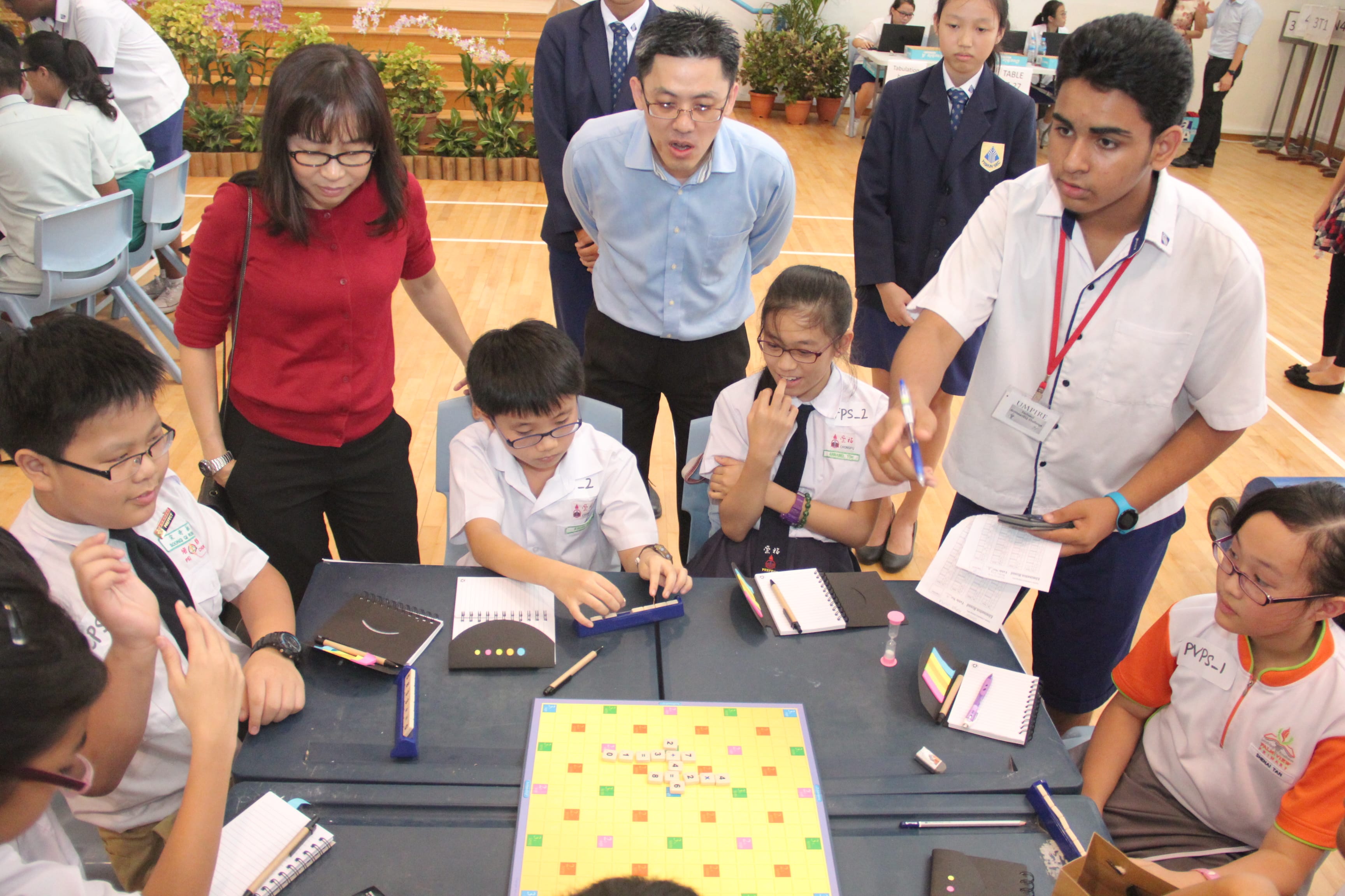Students and adults gather around a table with a colorful board game during a school competition event.