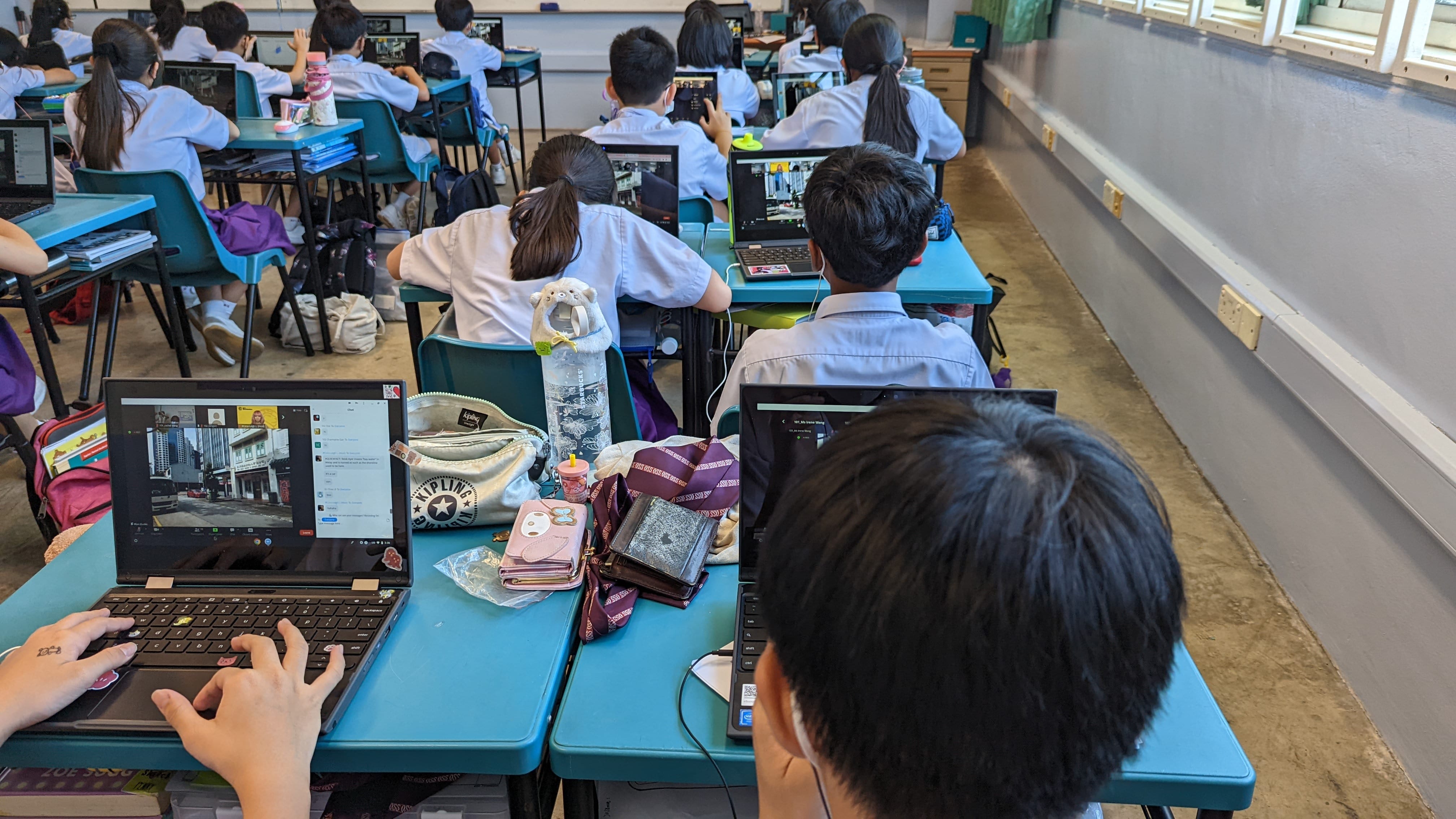 Students in white uniforms sit at teal desks using laptops in a full classroom.