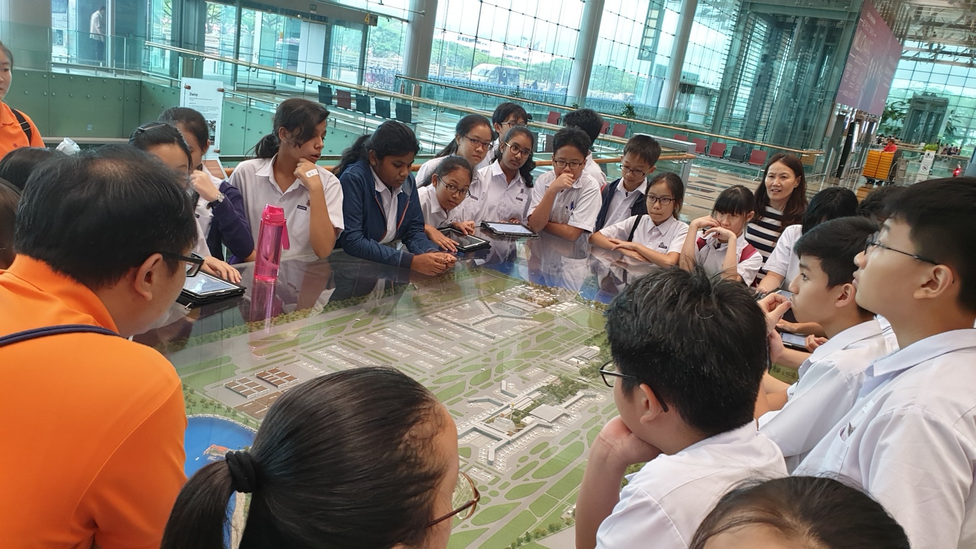 Students in white uniforms gather around a large architectural scale model inside a modern building with glass walls.