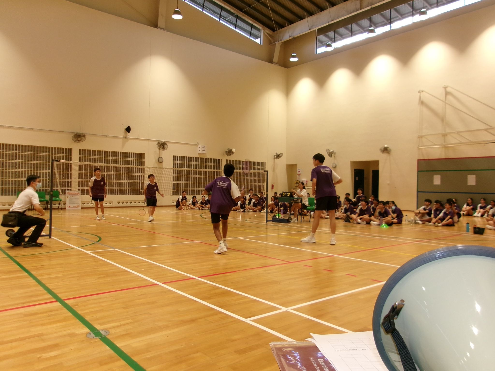 A group of students playing badminton in the Indoor School Hall supervised by a teacher.
