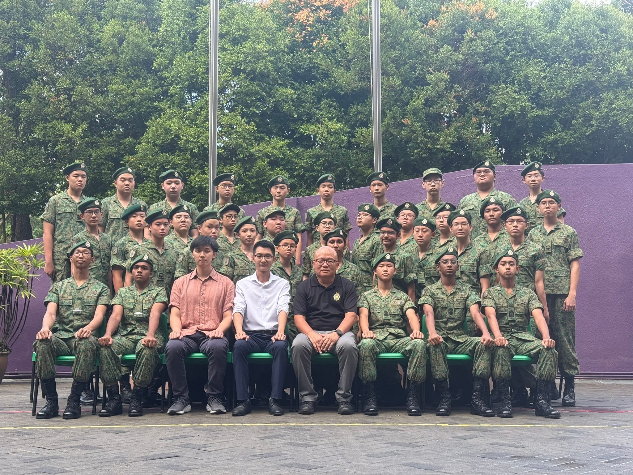 Large group of young people in green camouflage uniforms and berets posing with three adults in civilian clothes outdoors.