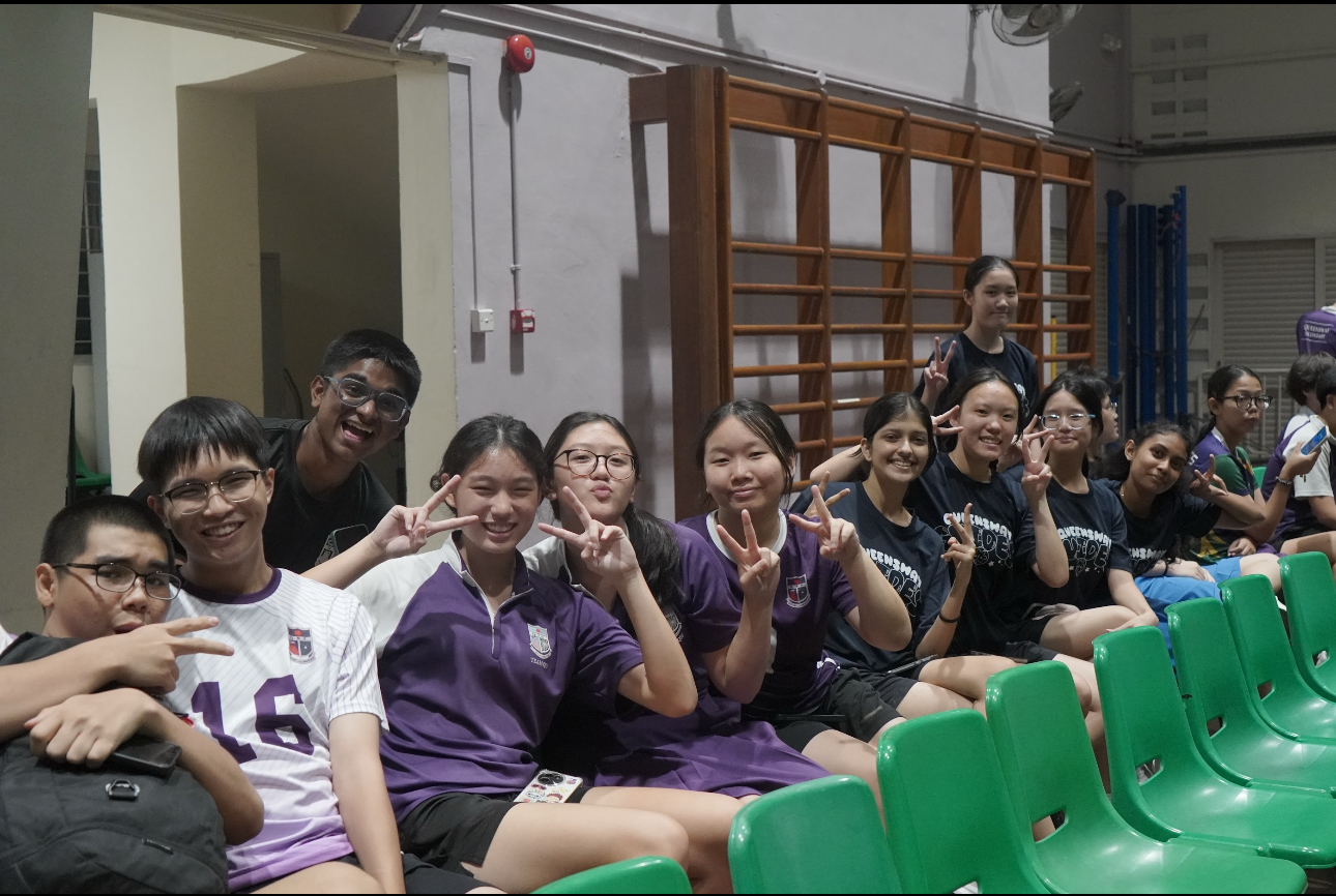 A group of smiling students in purple uniforms sit on green chairs in a gym, posing with peace signs.