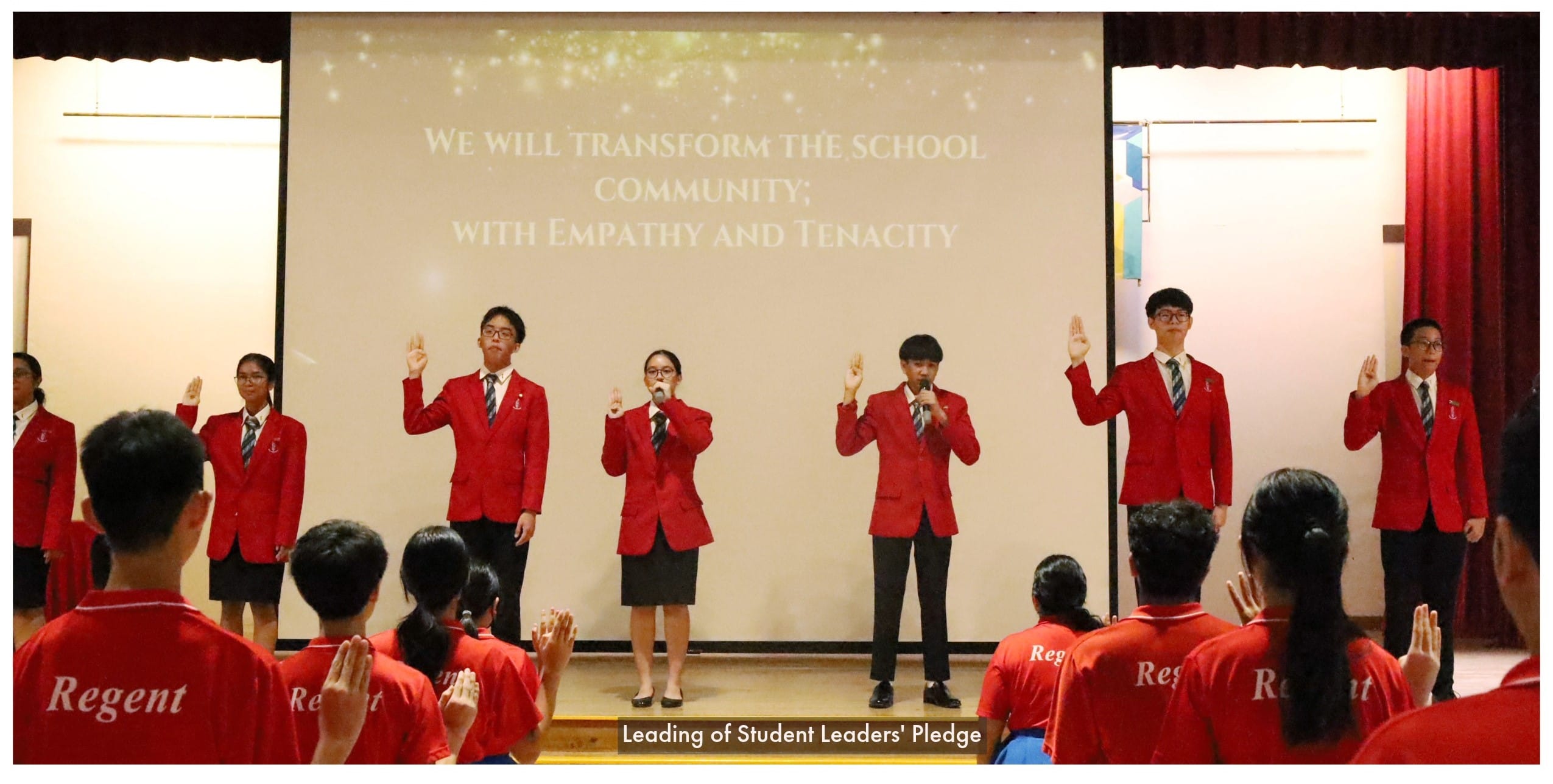 Students in red blazers lead a pledge on stage while seated students in red Regent shirts raise their hands.