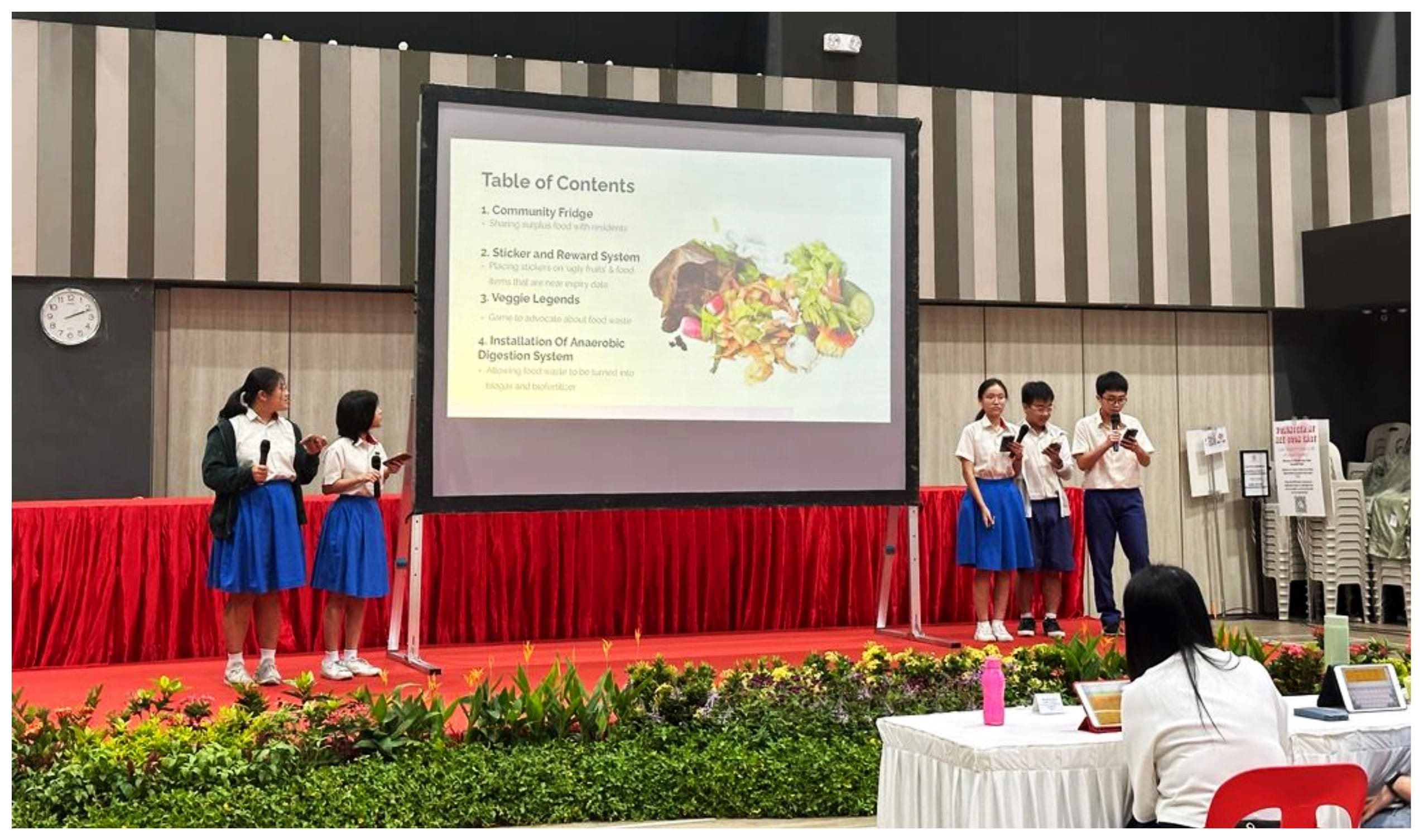 Four students in school uniforms present on stage with a slideshow titled Table of Contents about food waste solutions.