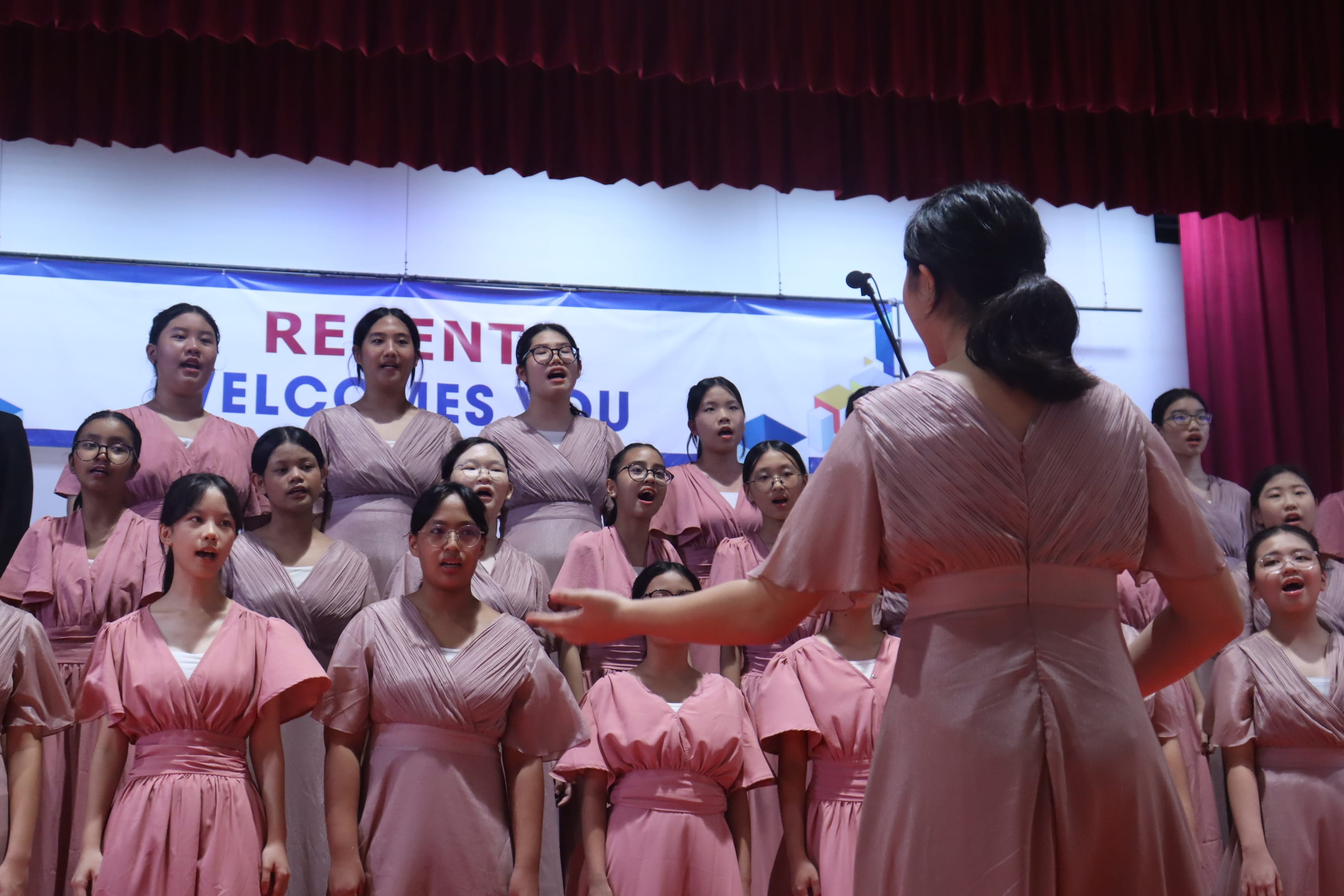 A choir of girls in pink dresses singing on stage, led by a conductor, with a banner reading "Regent Welcomes You."