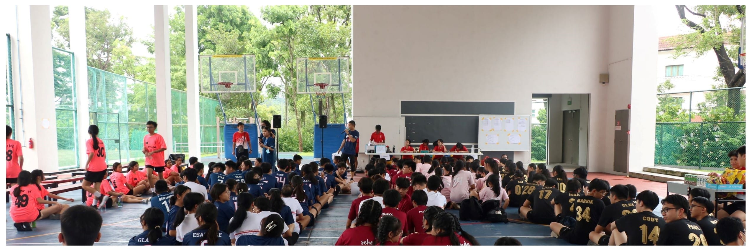 Students in pink, navy, red, and black jerseys sit on a gym floor during a school sports event briefing.